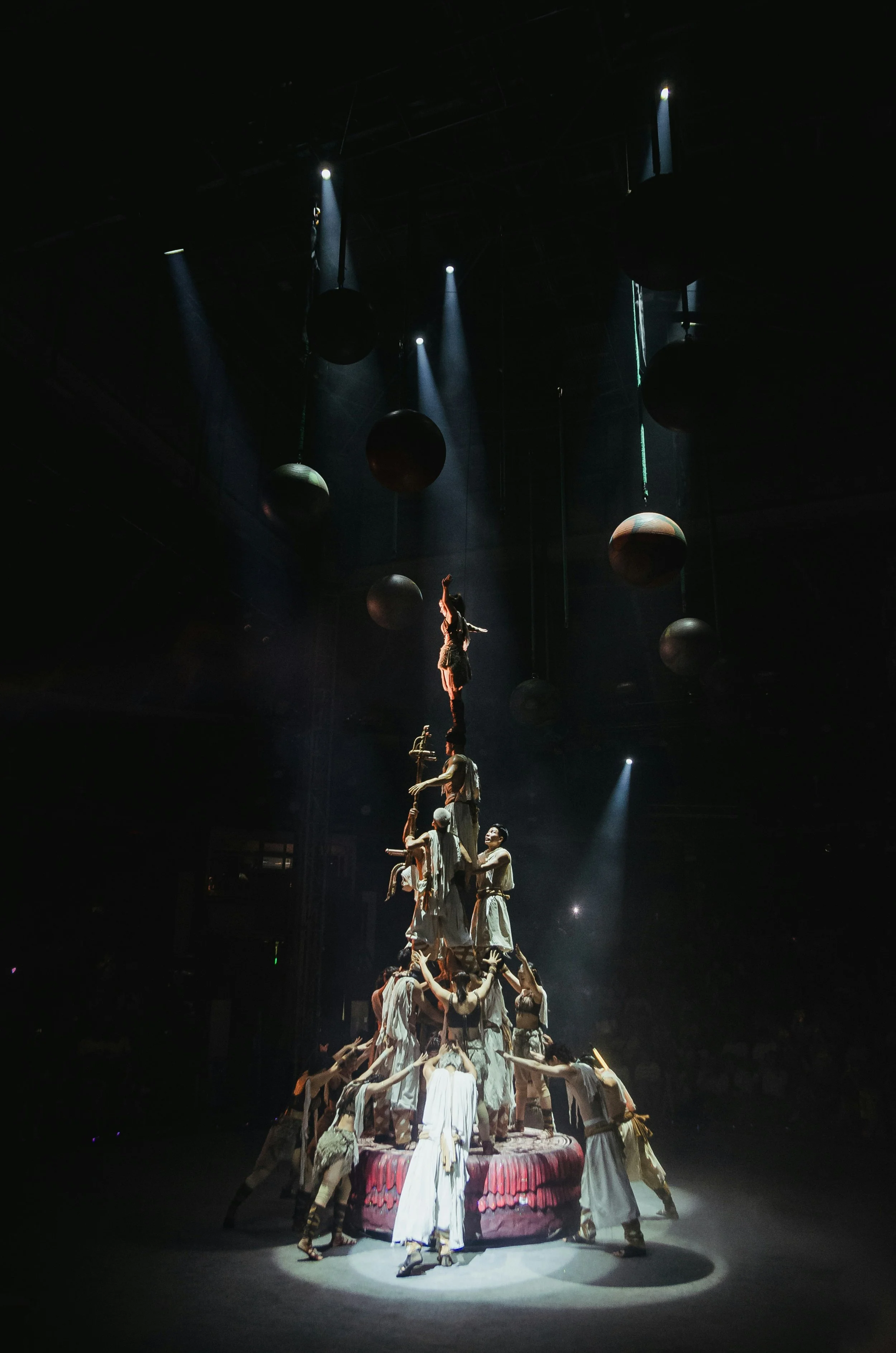A theatrical performance with performers forming a human pyramid on a circular platform, with some performers balancing on the structure and others reaching out towards it, under dramatic spotlights with hanging black spheres in a dark theater.