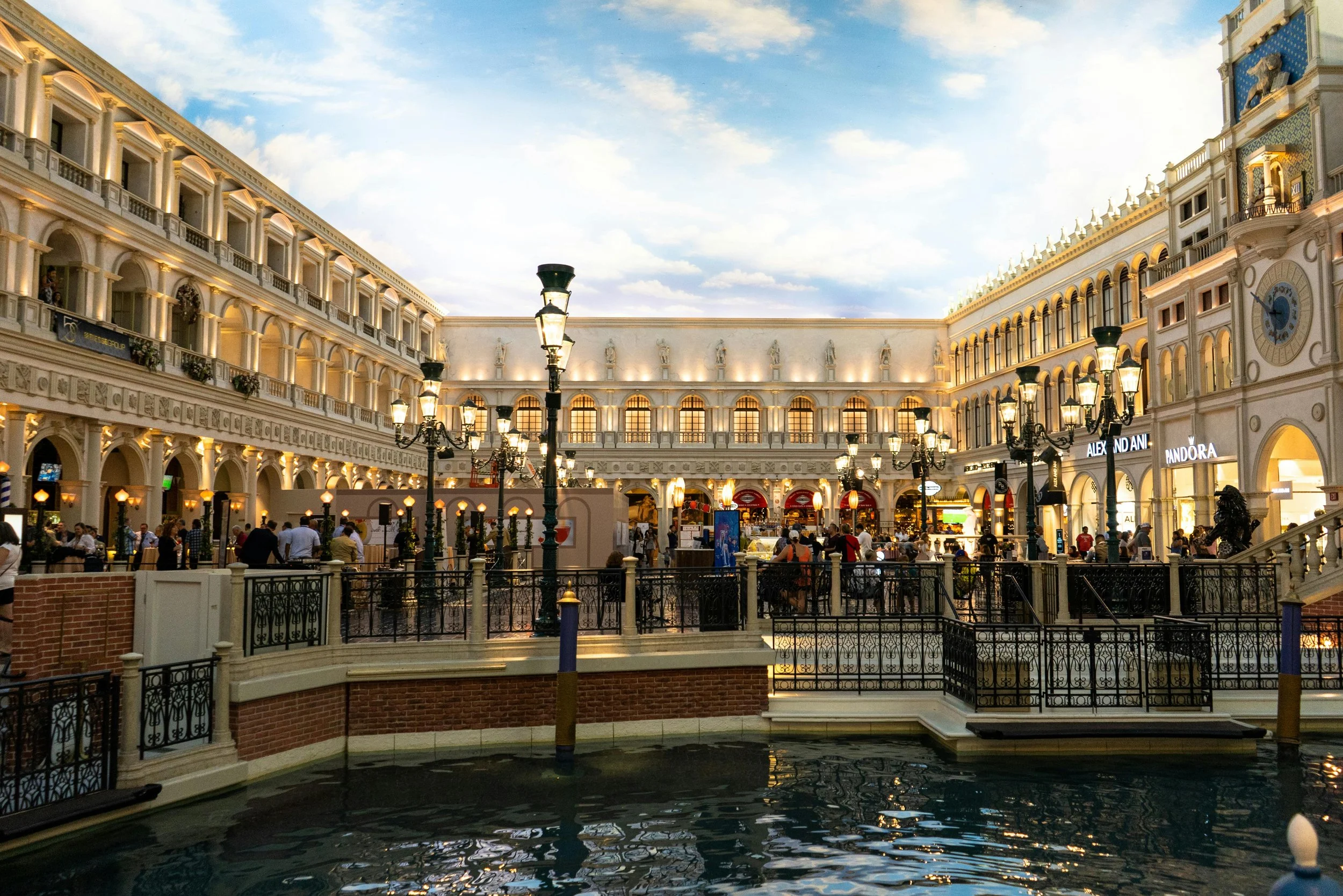 Indoor shopping mall with European-style architecture, decorative lamp posts, and a water feature in the foreground. Stores like Pandora are visible, and people are walking and shopping.