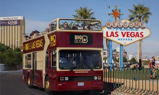 A red double-decker bus with tourists on top, parked near the iconic Vegas sign with palm trees, in Las Vegas, Nevada.