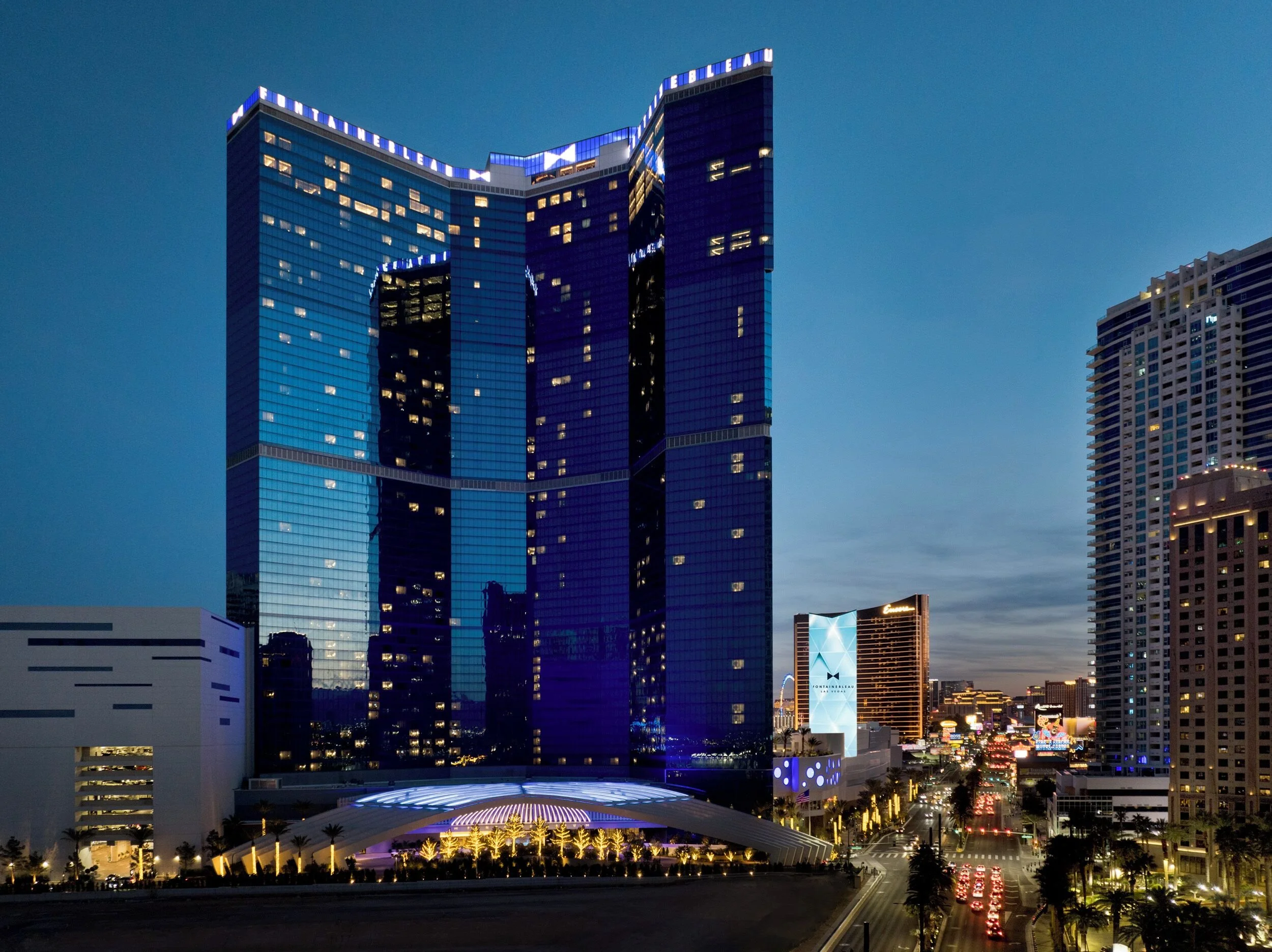 Nighttime cityscape with tall illuminated buildings, including a prominent blue glass skyscraper with lighting accents, and a busy street with red taillights and palm trees along the road.