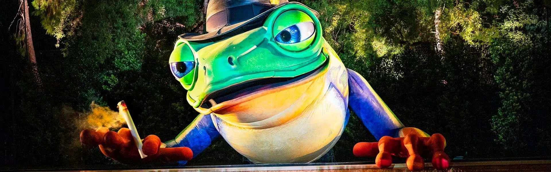 Colorful giant frog sculpture with glasses, holding a cigarette, illuminated at night, surrounded by trees.