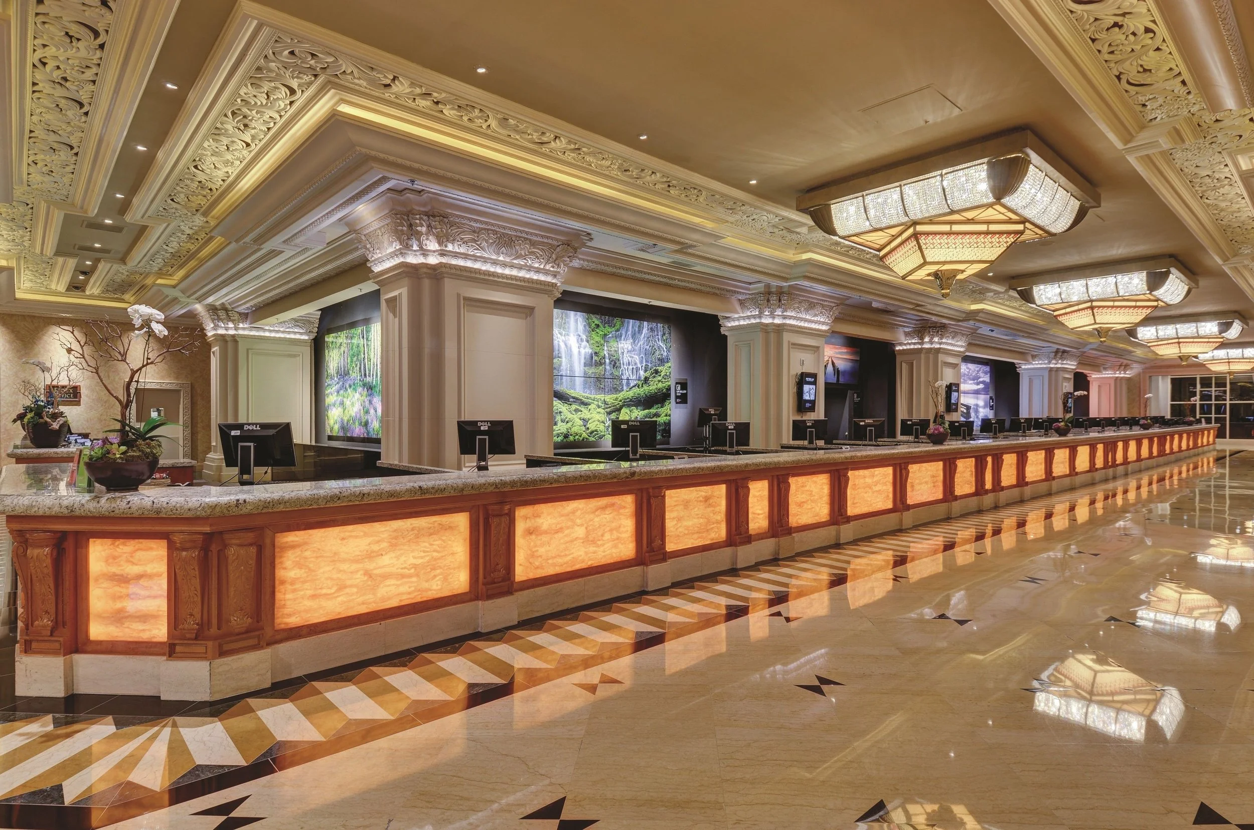 Front desk reception area with multiple check-in counters, ornate ceiling with large chandeliers, framed landscape screens behind the counters, and polished marble floors.