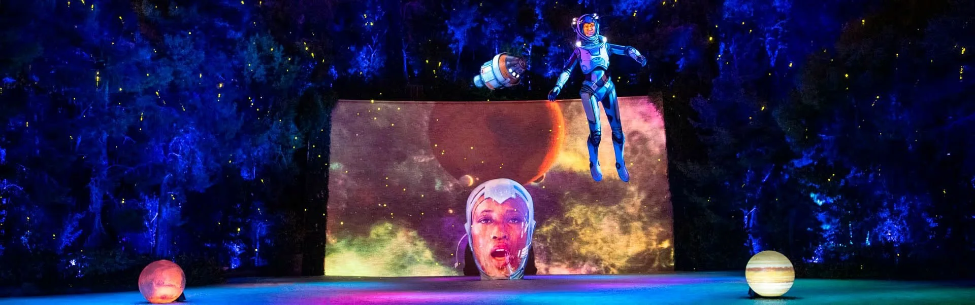 Stage scene with two characters in space-themed costumes, one floating and one in front of a large screen displaying a woman's face and space imagery, with two planet models on the stage.