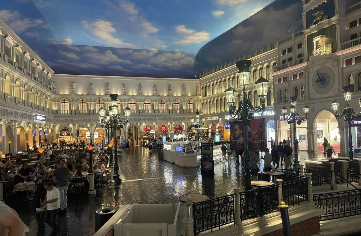Indoor shopping mall designed to look like a European city with a painted sky ceiling, ornate architecture, and street lamps, filled with people dining and shopping.