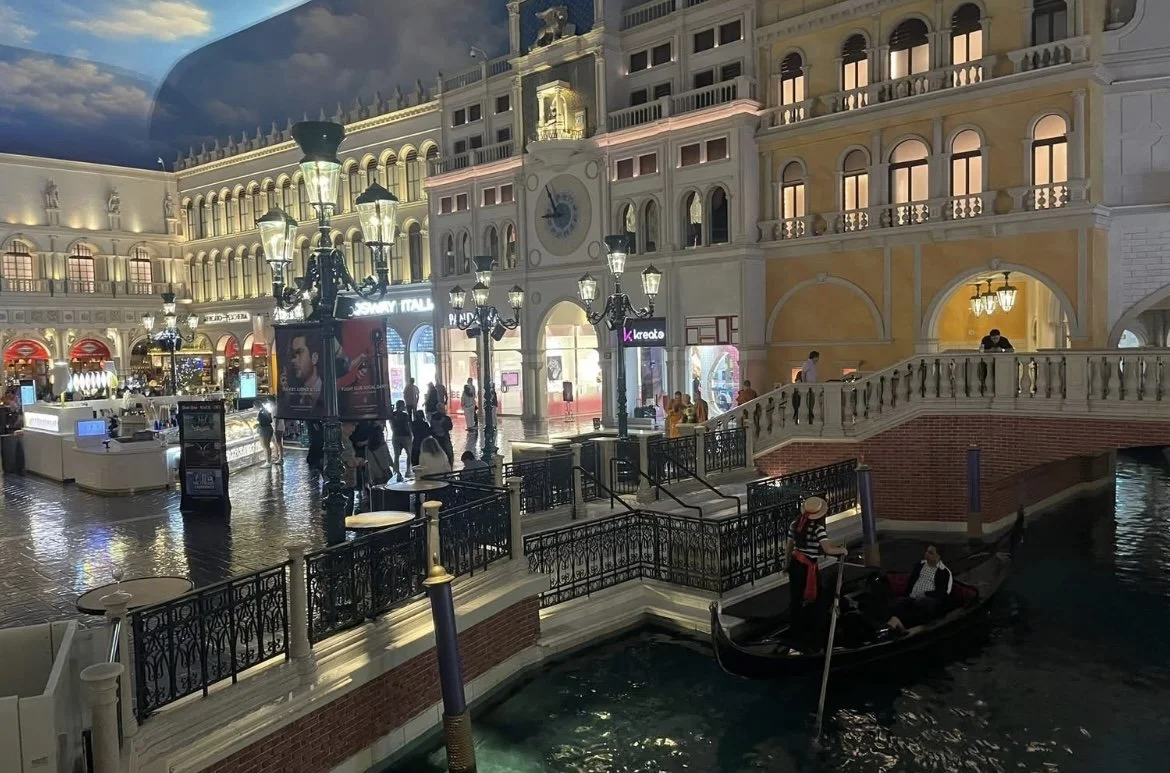 Indoor shopping mall with European-style architecture, canals with gondolas, and people walking and shopping.