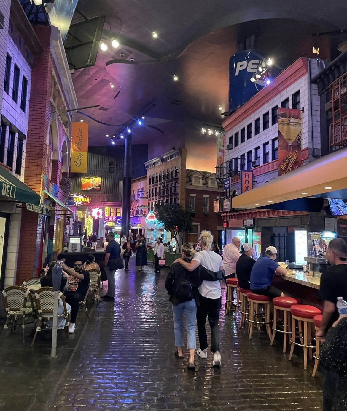 Indoor entertainment district with neon lights, bar seating, and people walking and socializing.