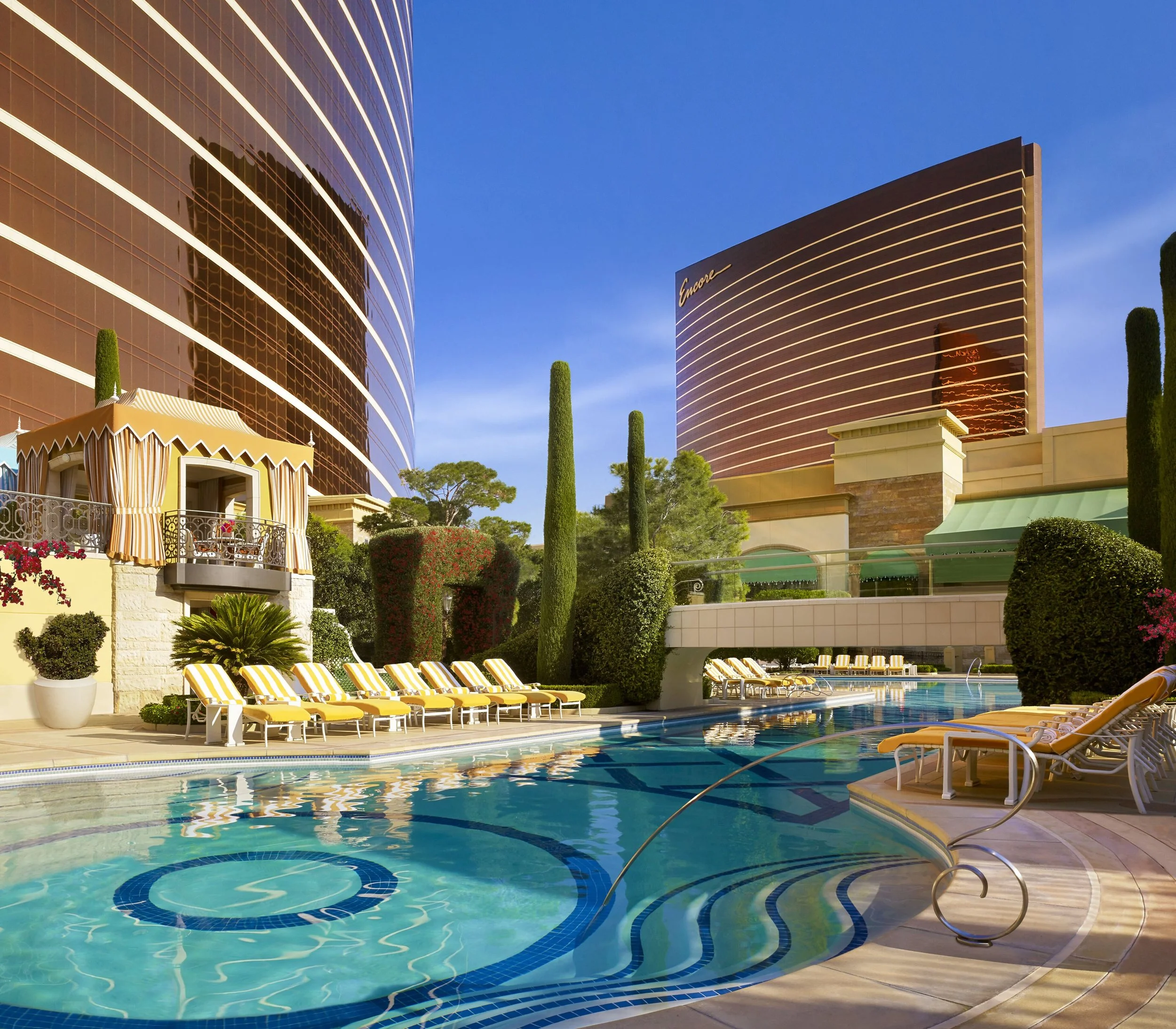 Luxury hotel rooftop swimming pool area with yellow lounge chairs, cabanas, tall green cypress trees, and modern high-rise buildings in the background under a clear blue sky.
