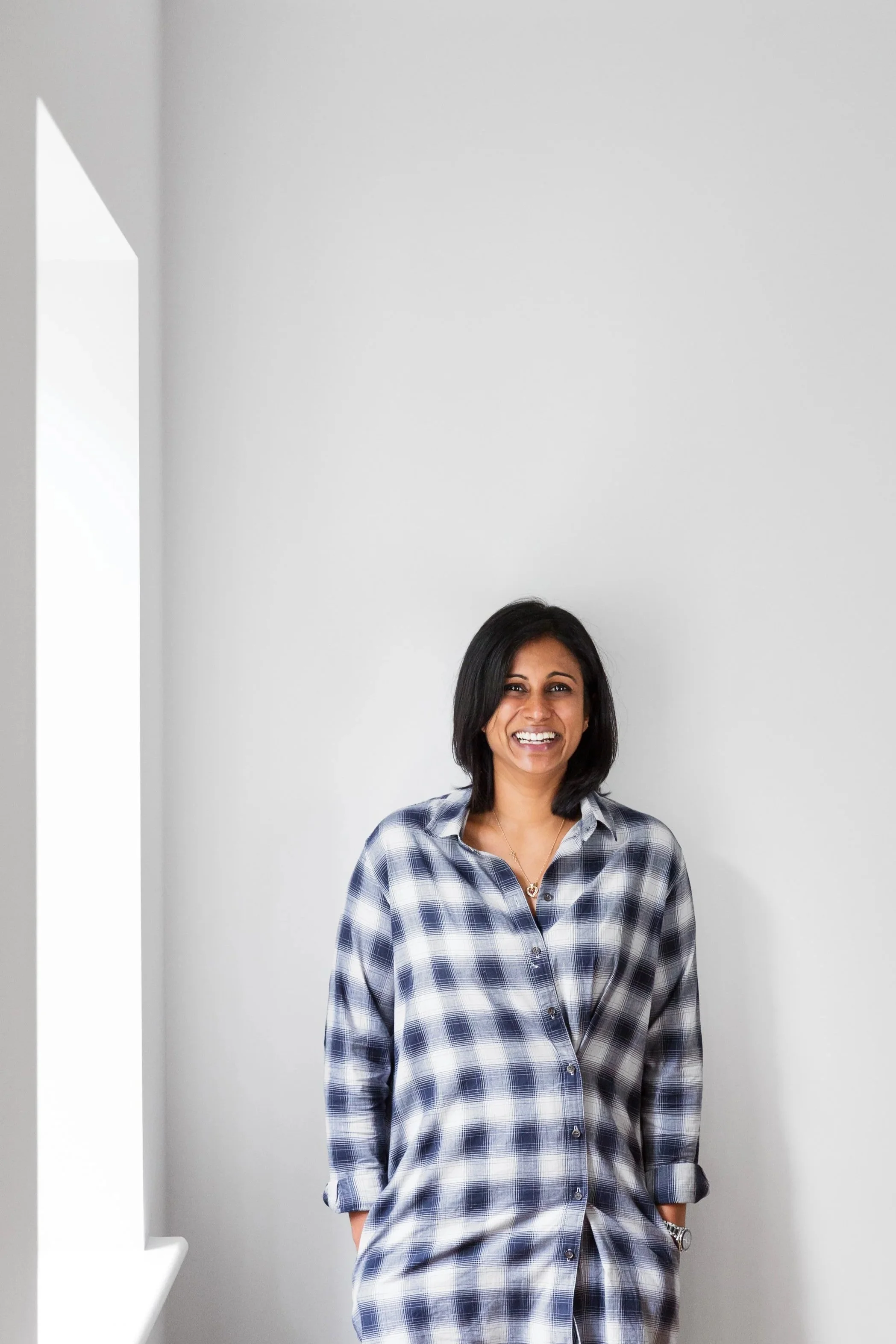 A woman with black hair smiling and wearing a blue and white plaid shirt, standing against a plain white wall near a window.