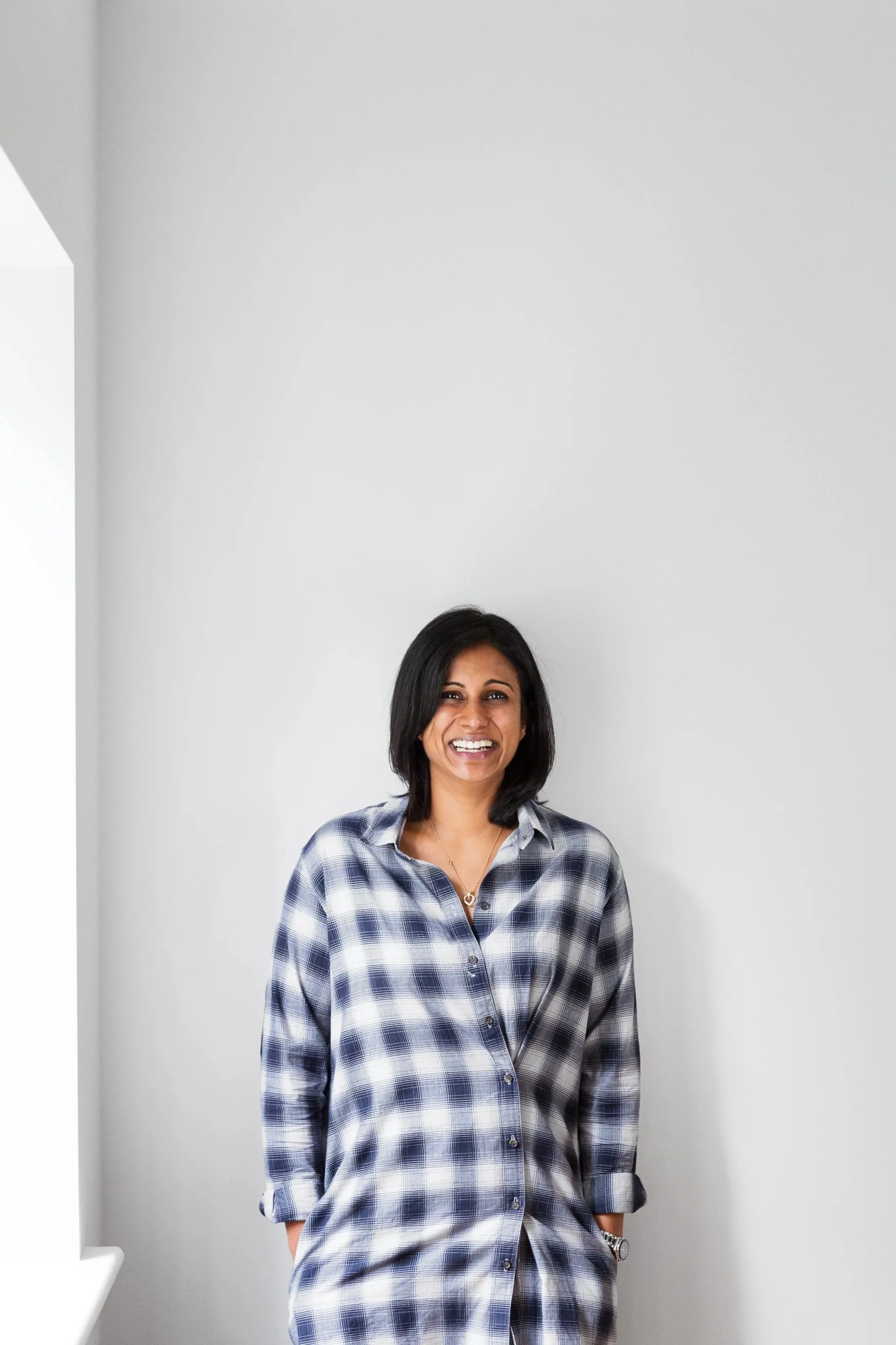 A woman with black hair smiling and standing against a plain white wall, wearing a blue and white plaid shirt and a wristwatch.