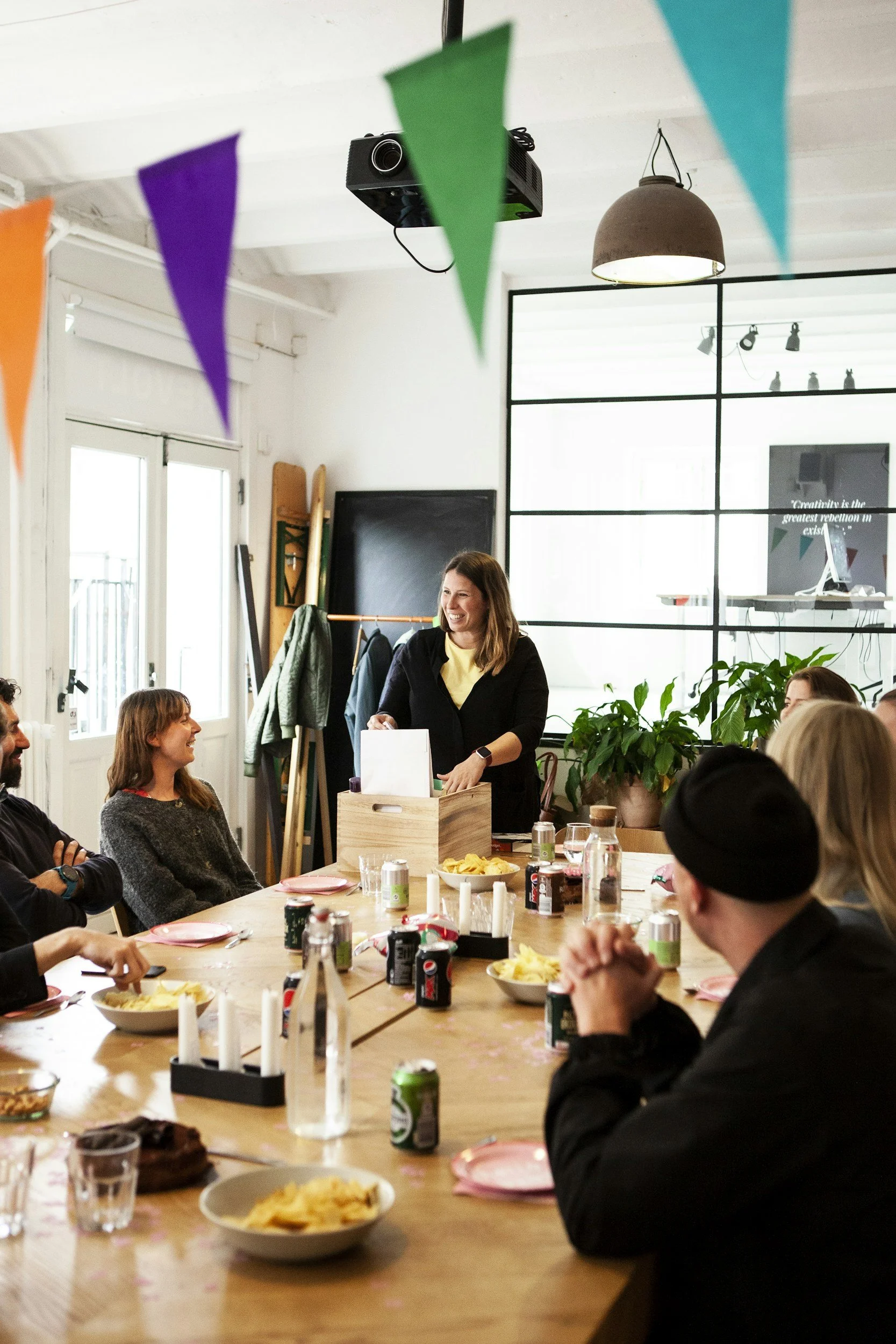 People gathered around a large dining table in a bright, modern room, celebrating an event with food and drinks. A woman is standing and smiling while handing something to a guest. The room is decorated with colorful bunting, and there is a large window and plants in the background.