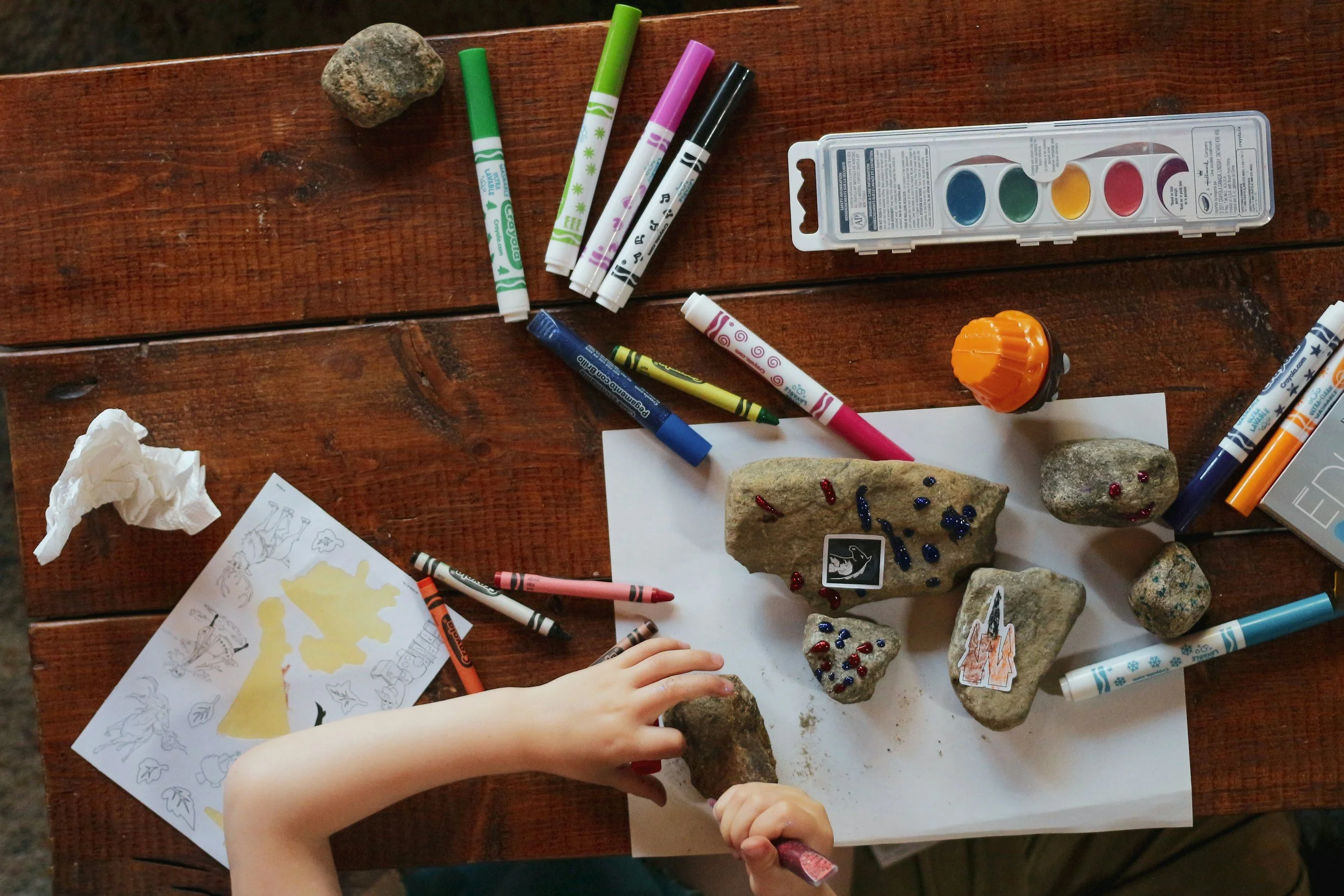 Child's hand arranging rocks on a table with paint, markers, stickers, and coloring sheets, creating a craft project.