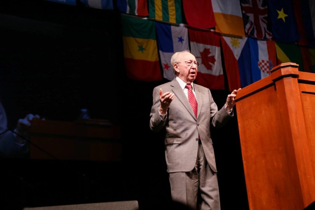 An elderly man in a gray suit and red tie speaking at a podium, with international flags hanging behind him.