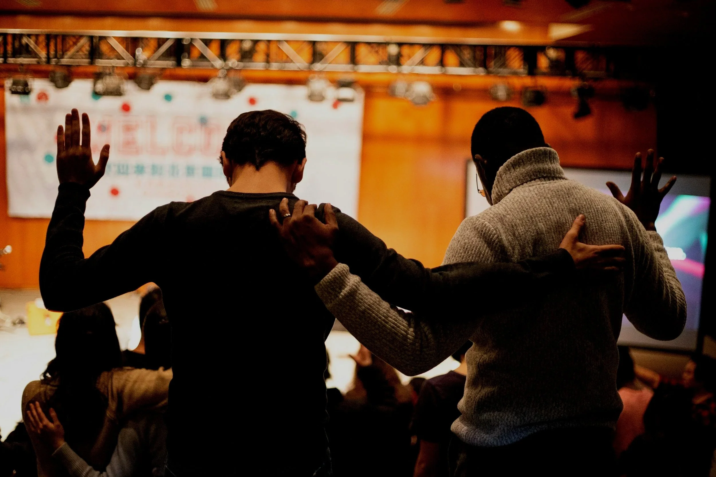 Two people in an indoor setting with wooden walls and stage lighting, standing close and holding each other's hands and arms in a gesture of prayer or reflection, with a group of people sitting in the background.
