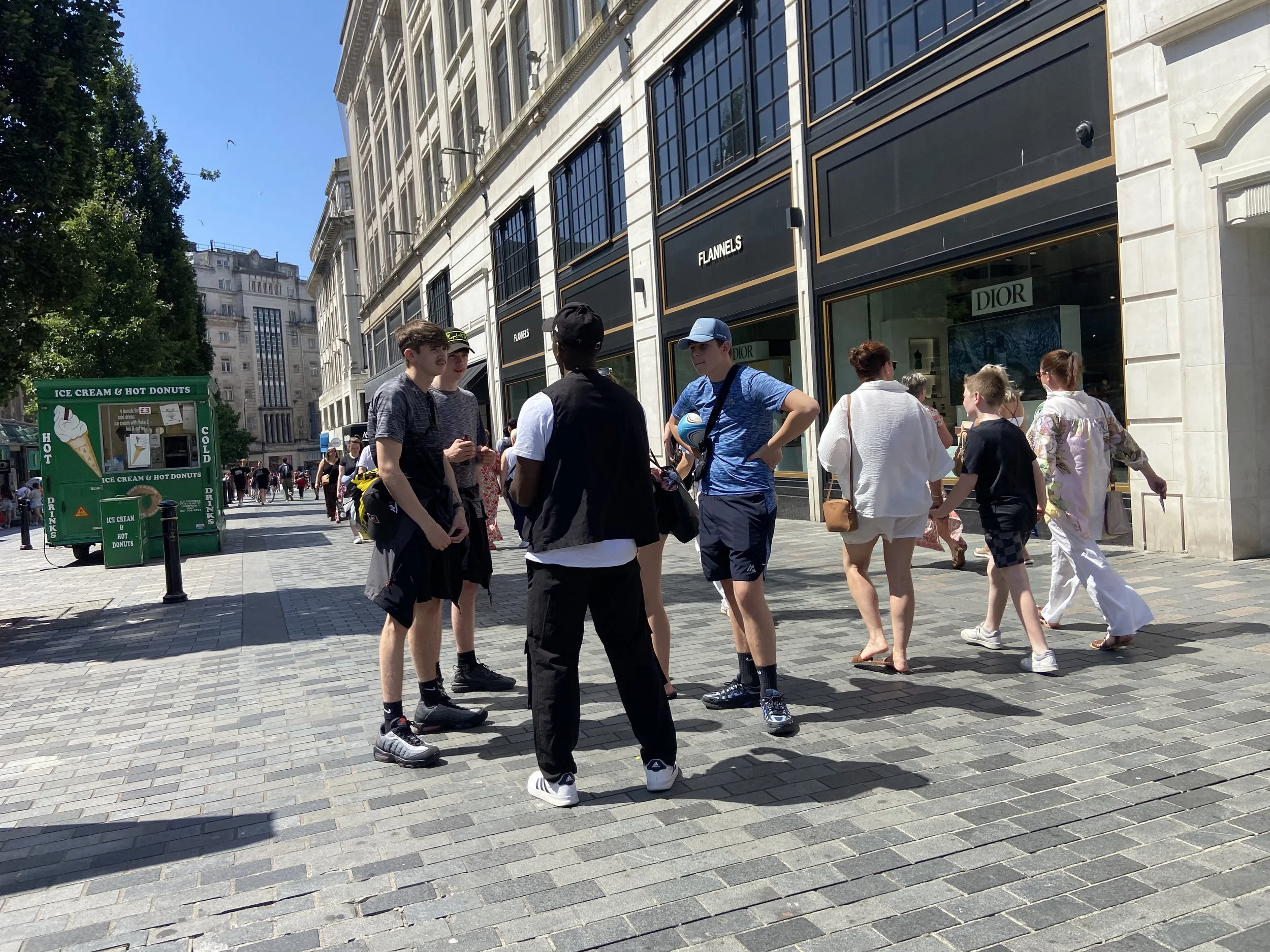A group of young people standing and talking on a busy city sidewalk with storefronts, trees, and a green ice cream stand in the background.