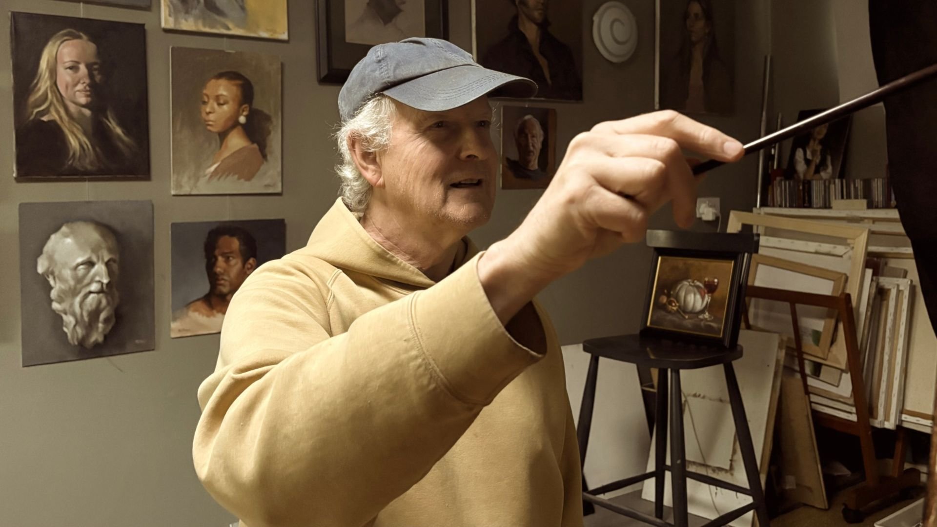 Simon Broom working on an oil painting at his easel, surrounded by portraits and plaster busts in his art studio