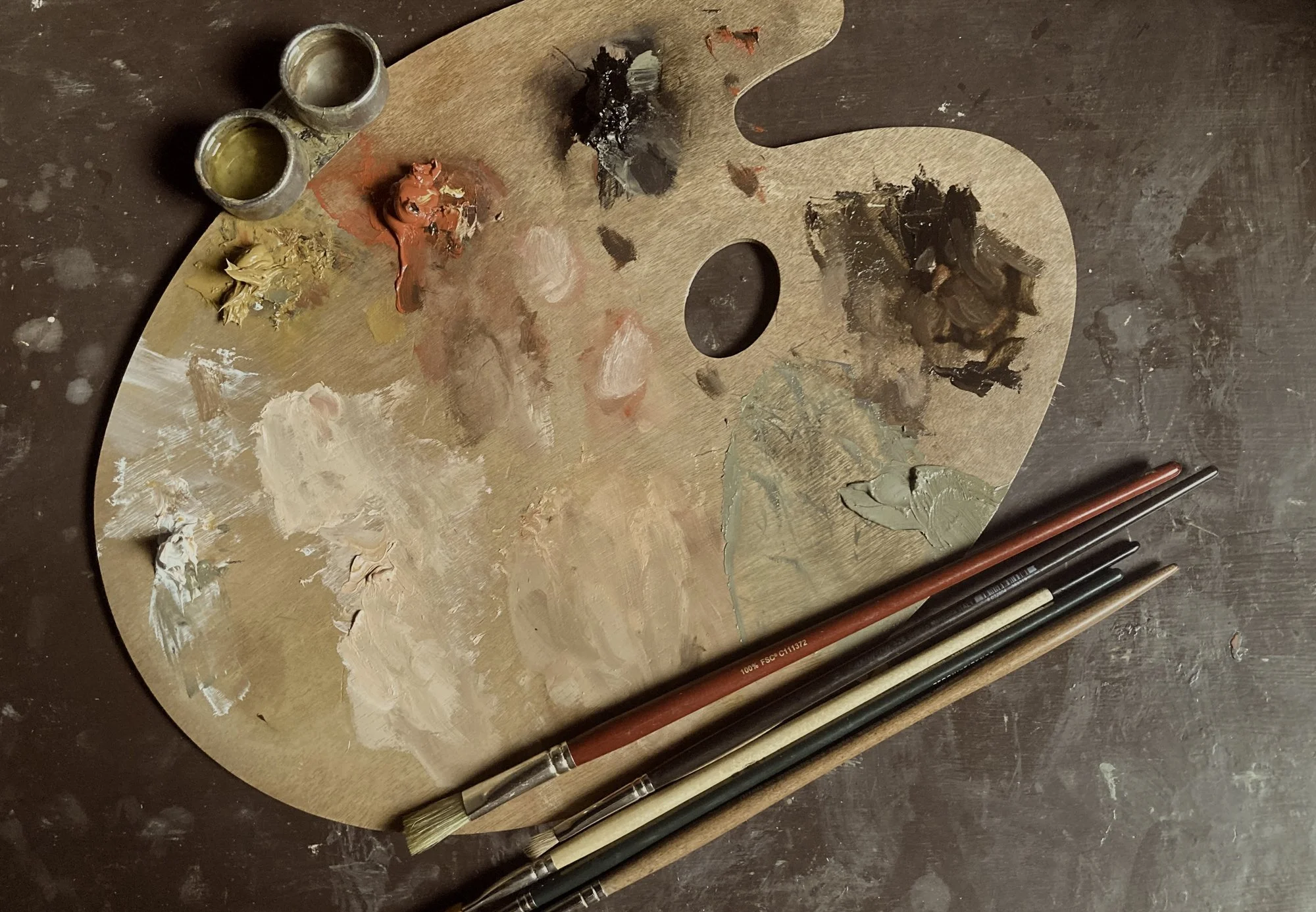 Wooden artist's palette with oil paints in yellow, red, black and white, and three paintbrushes laid below it