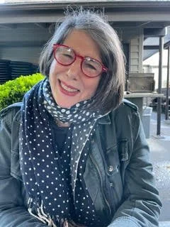 Smiling woman with gray hair and red glasses, wearing a blue polka dot scarf and leather jacket, sitting outdoors with a background of green foliage and industrial structures.