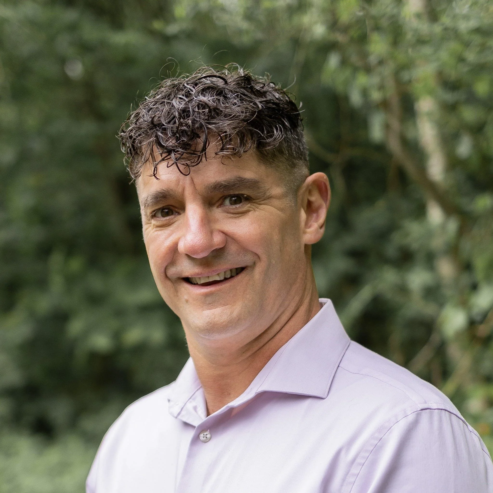 A man with curly dark hair, wearing a light purple button-up shirt, smiling outdoors with blurred green foliage in the background.