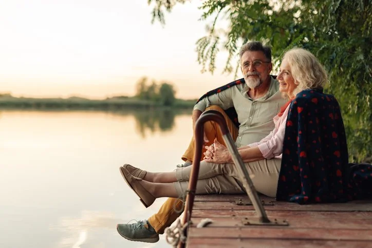 An elderly couple sitting on a dock by a lake at sunset, holding hands and enjoying each other's company.