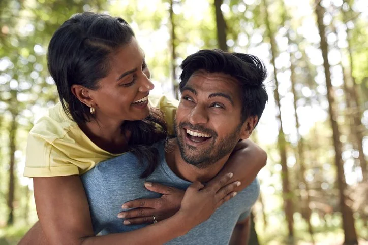 A woman gives a piggyback ride to a man in a forest with green trees, both smiling and enjoying each other's company.