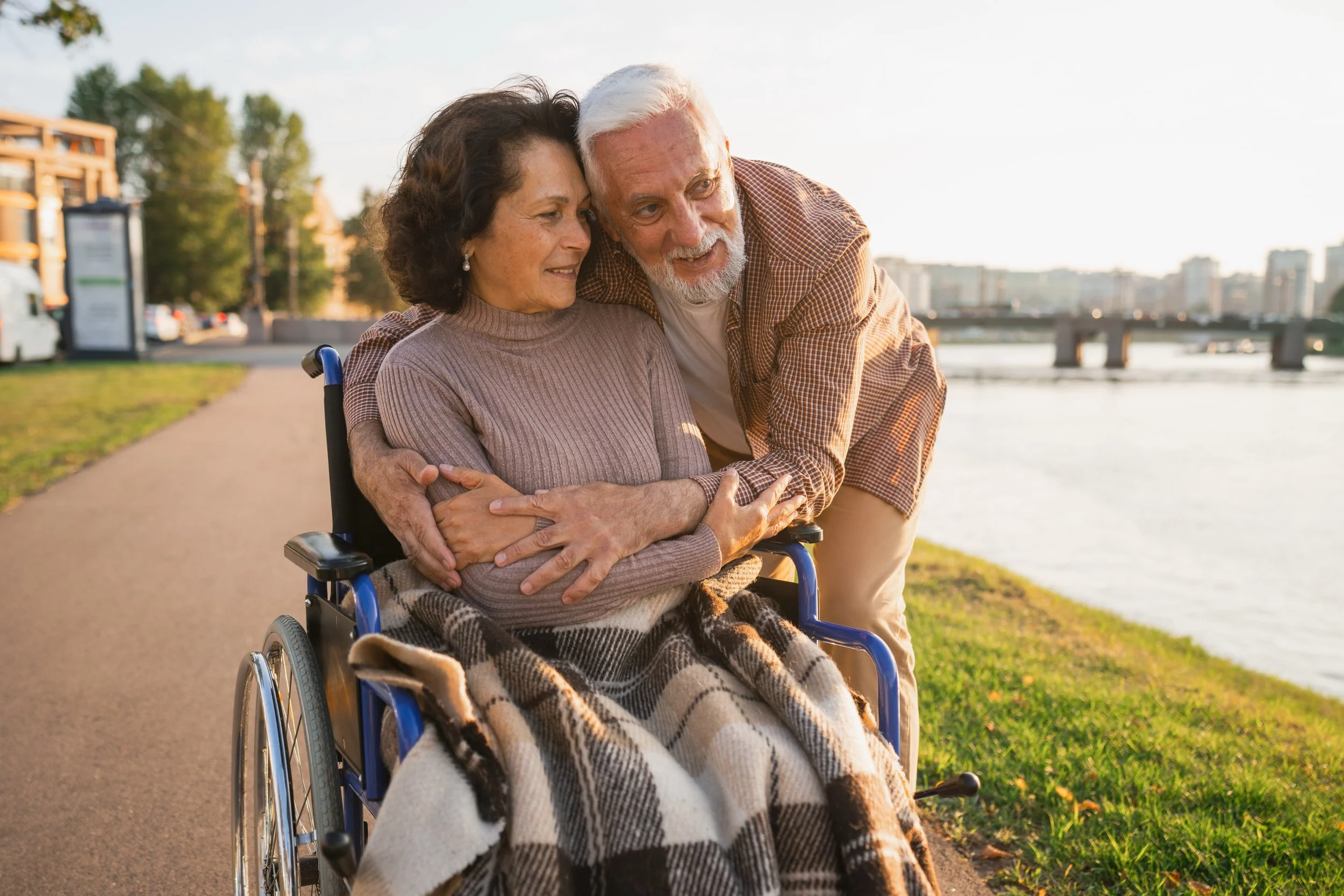 An elderly woman sitting in a wheelchair and an elderly man hugging her outdoors near a river with a city skyline in the background during sunset.