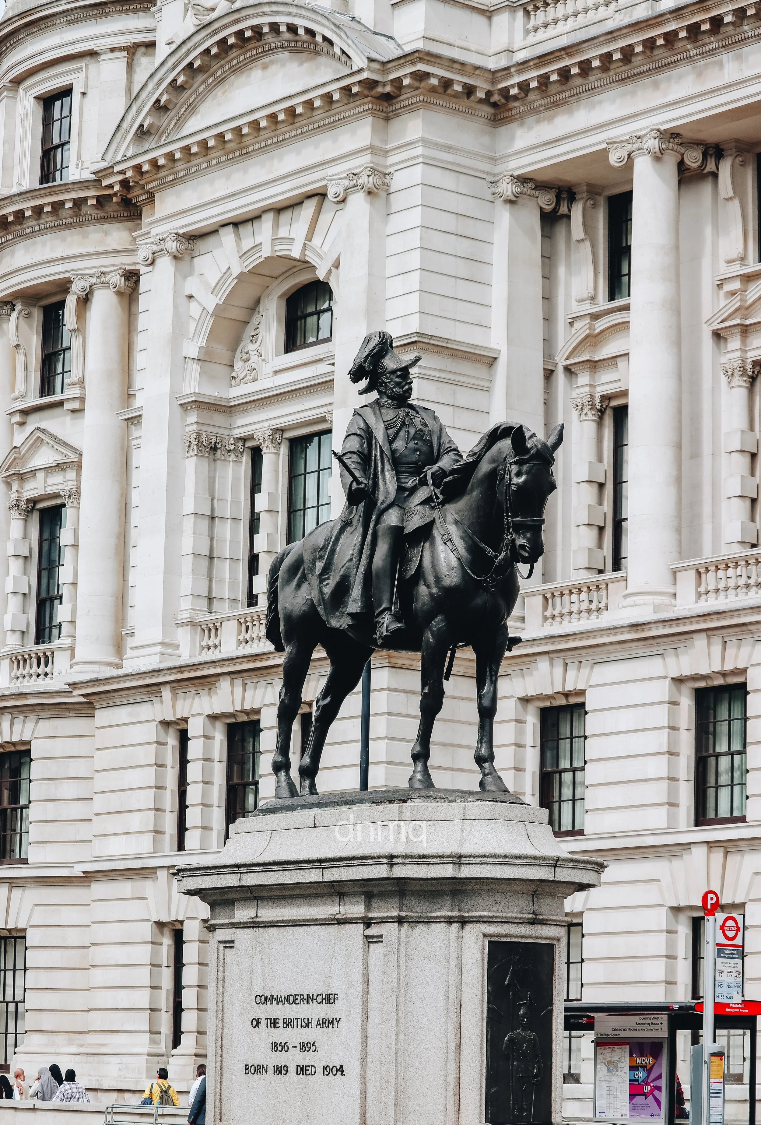 Bronze statue of a man on horseback located in front of a historic white stone building, with people walking nearby and a red and white bus stop sign in the background.