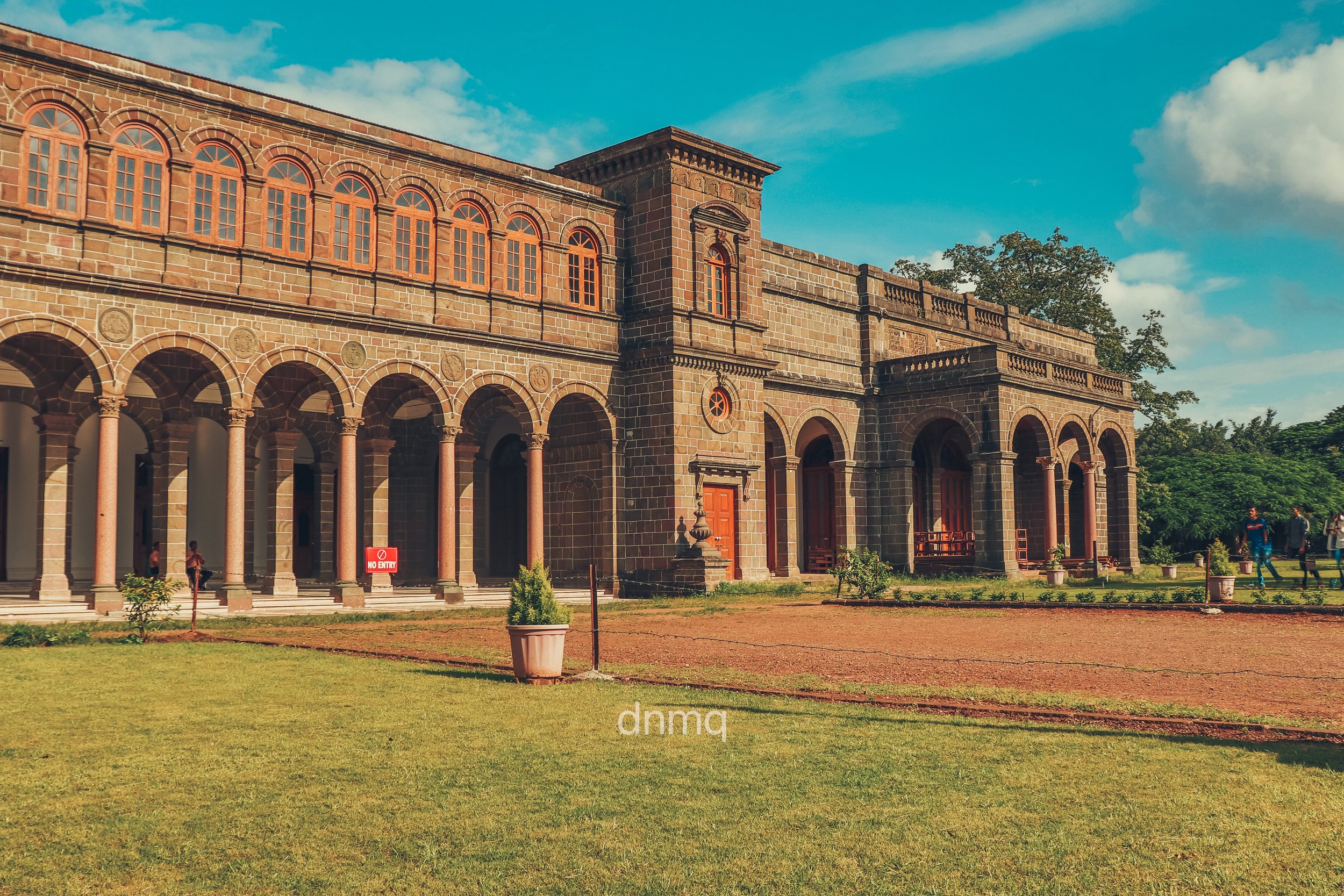 Historic stone building with arched windows and columns, green lawn in the foreground, and people walking outside under a partly cloudy sky.
