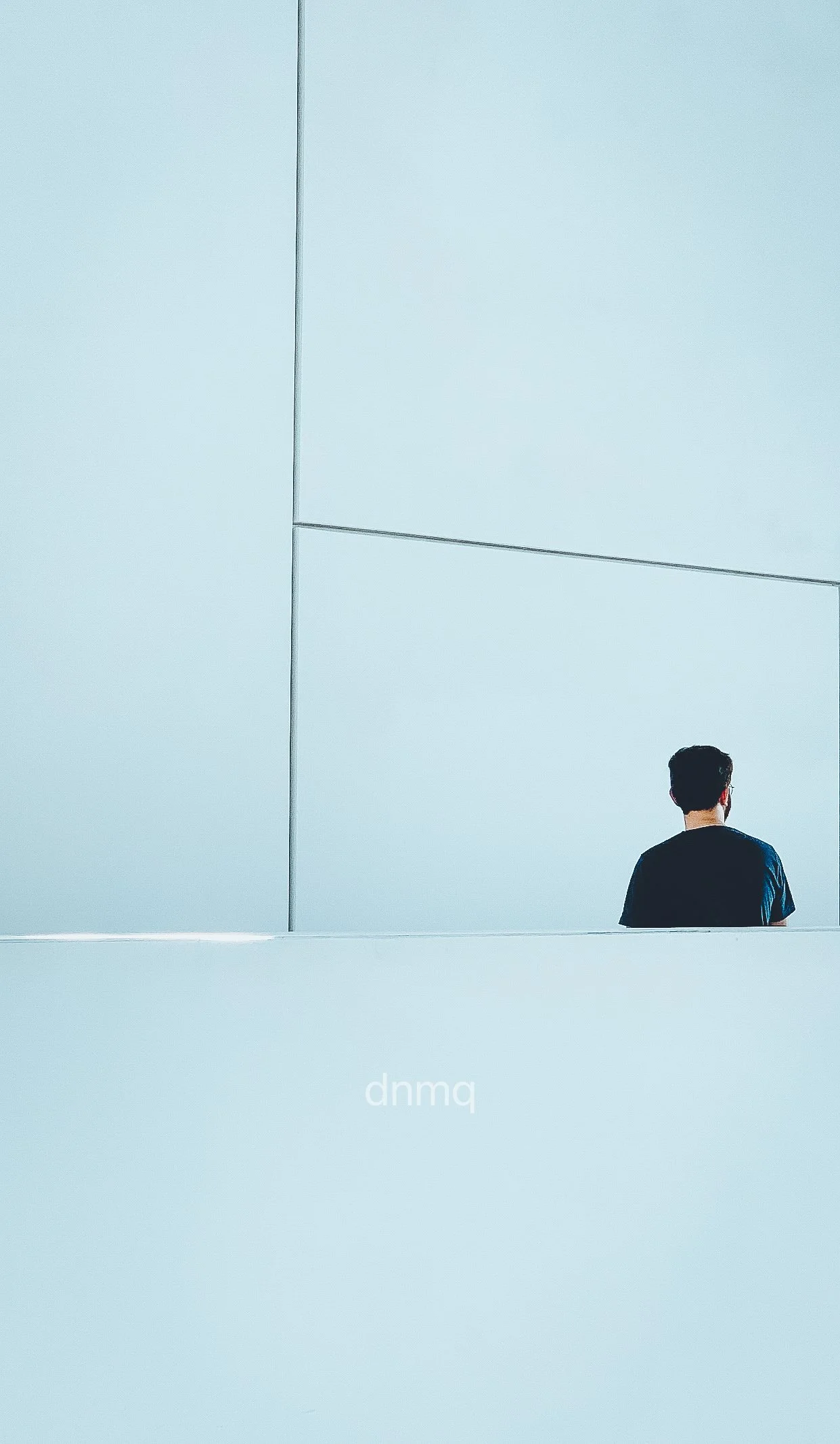 A person with dark hair wearing a black shirt, looking at a large minimalist pale blue wall with geometric lines.