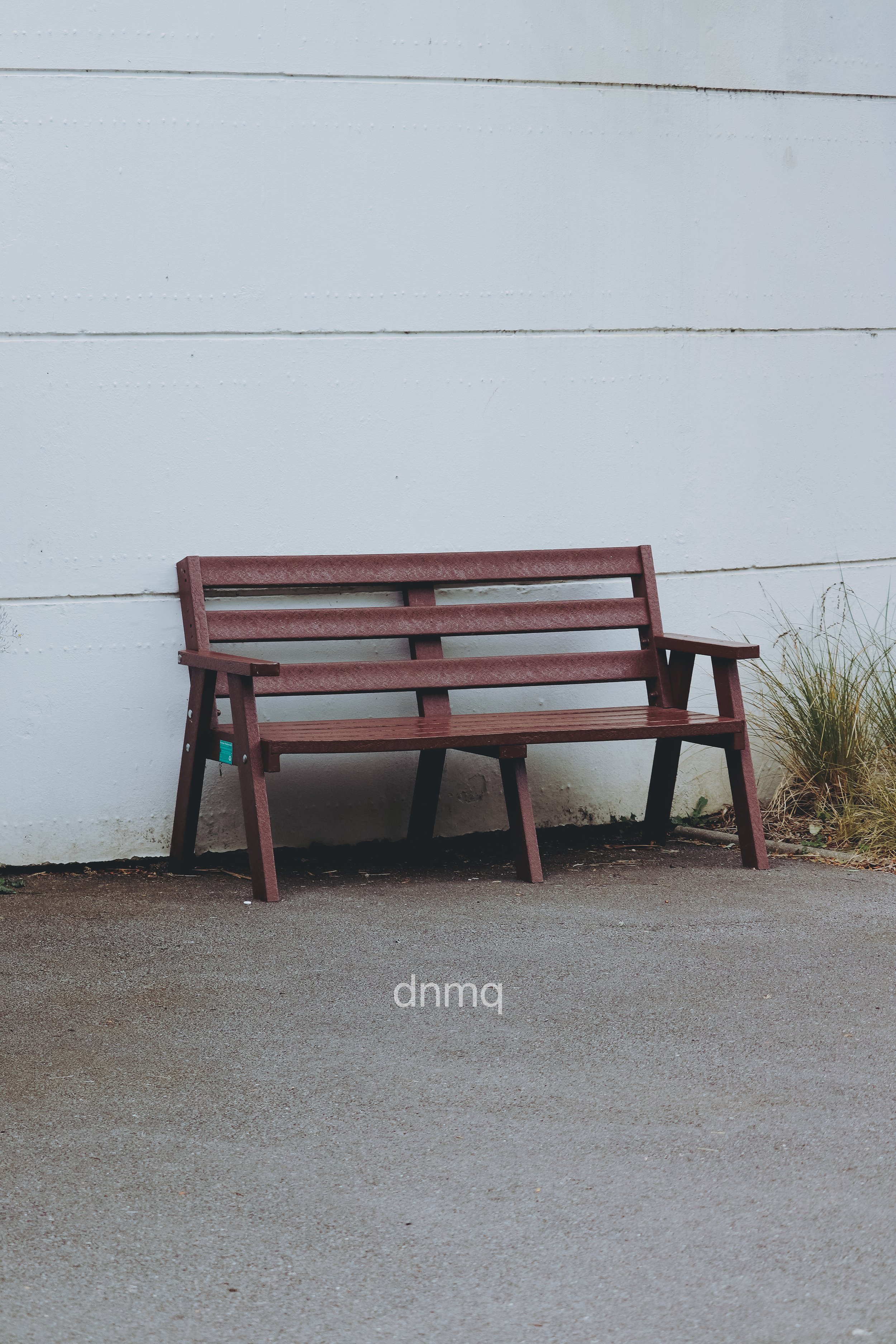 A red wooden park bench placed against a white wall, with grass on the right side.