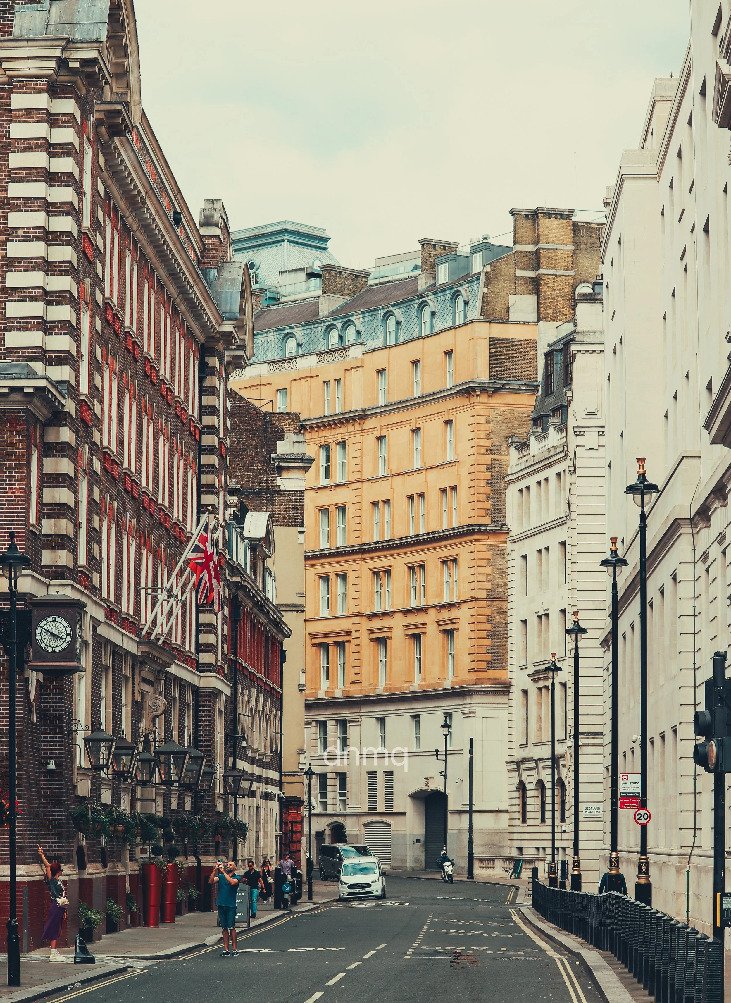 A city street lined with historic buildings, some with brick and stone facades, and street lamps. People are walking on the sidewalk, and cars are on the road.