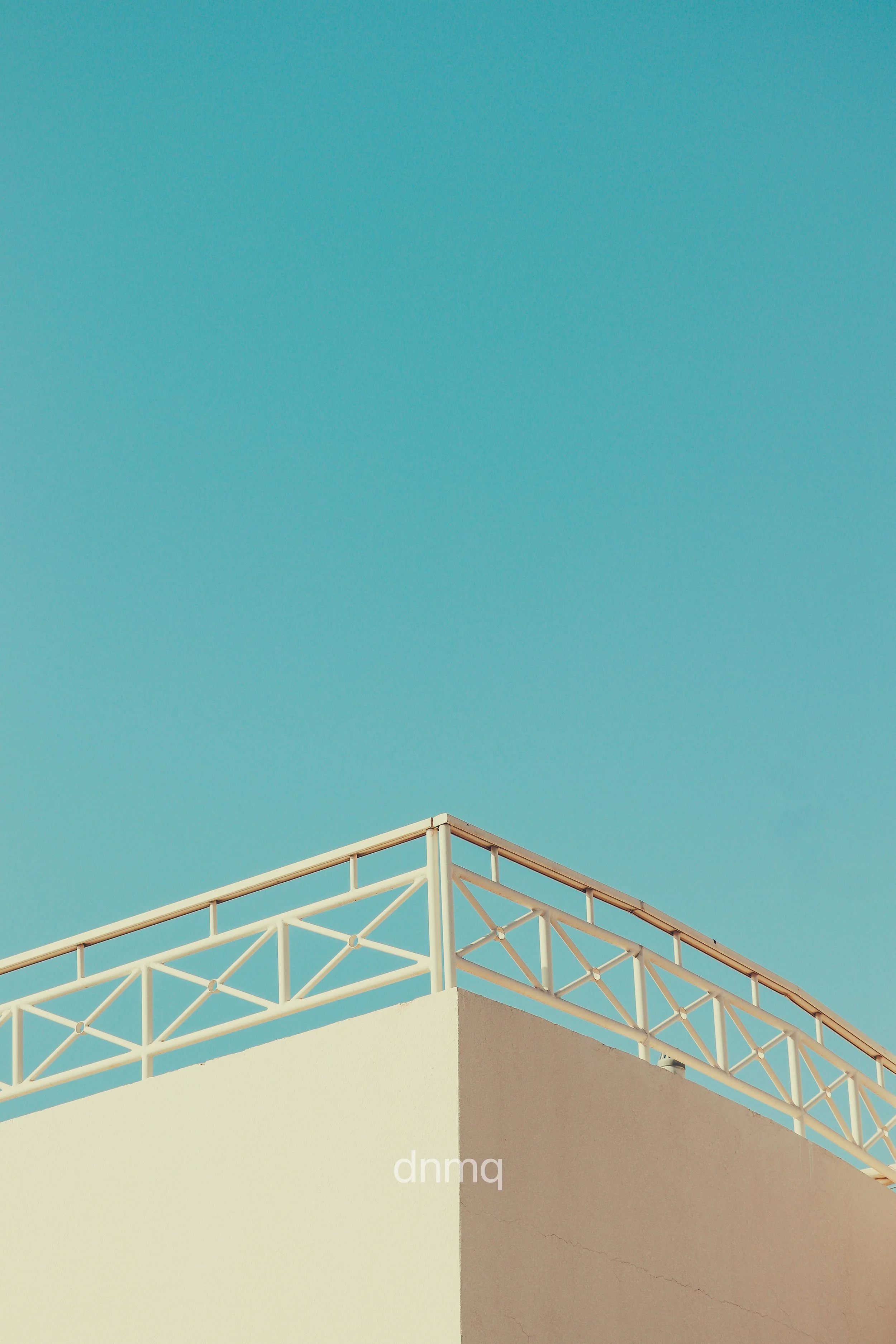 A building corner with a beige wall and white balcony railing against a clear blue sky.