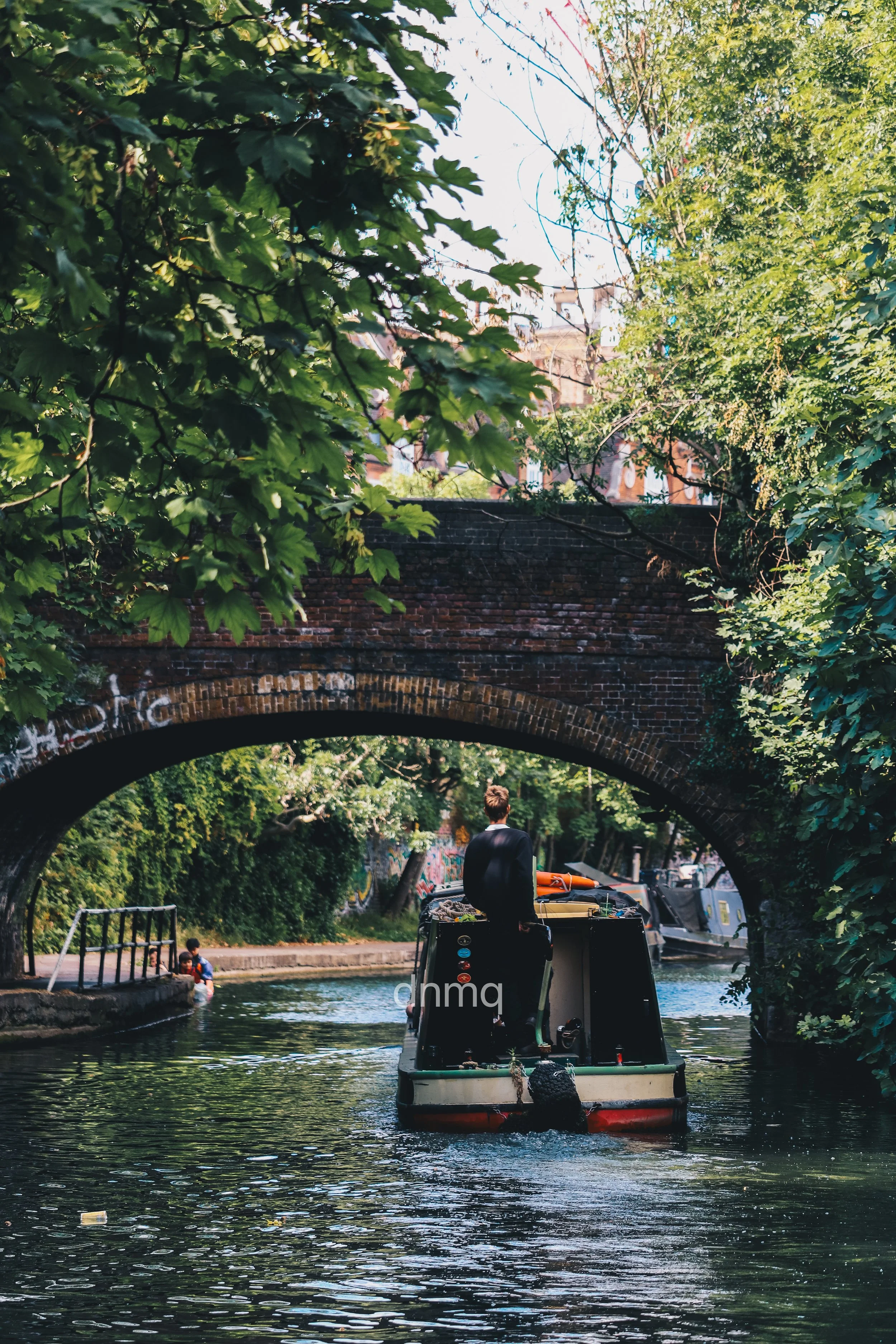 A person in a black jacket is on a boat on a narrow waterway, passing under an old brick bridge with greenery on either side. The scene is urban and peaceful with trees and buildings in the background.