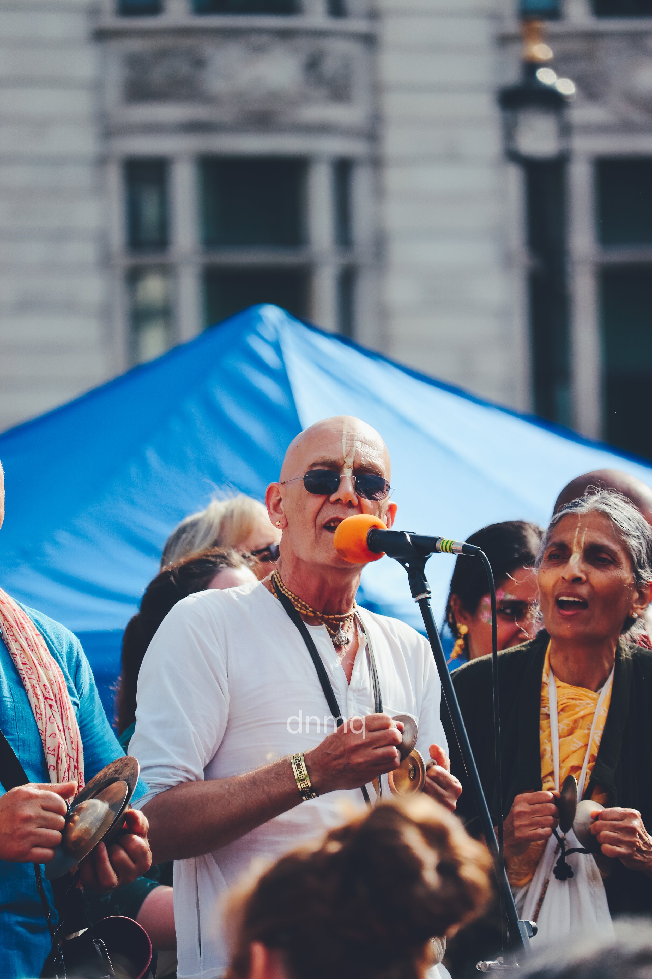 Man with a shaved head, sunglasses, and traditional Hindu markings on his forehead singing into a microphone at an outdoor gathering, with others around him and a blue tent in the background.