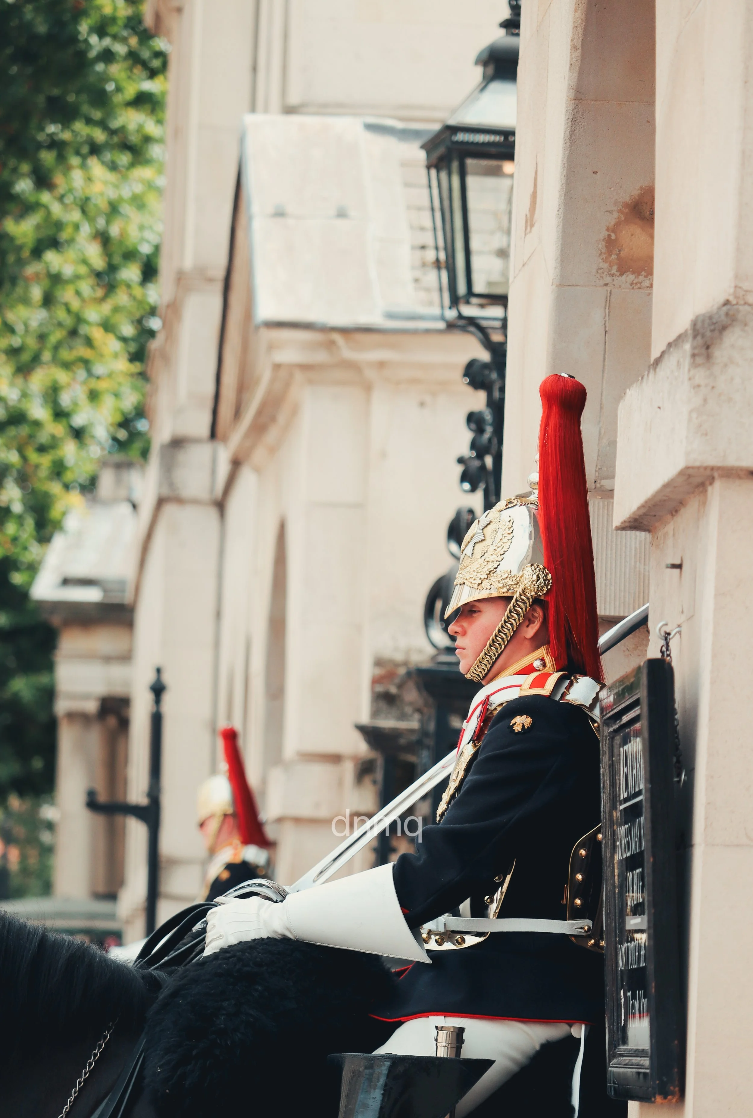 A member of a ceremonial guard in a decorated uniform with a red plume, sitting on a horse and leaning against a building, during a parade or ceremony.