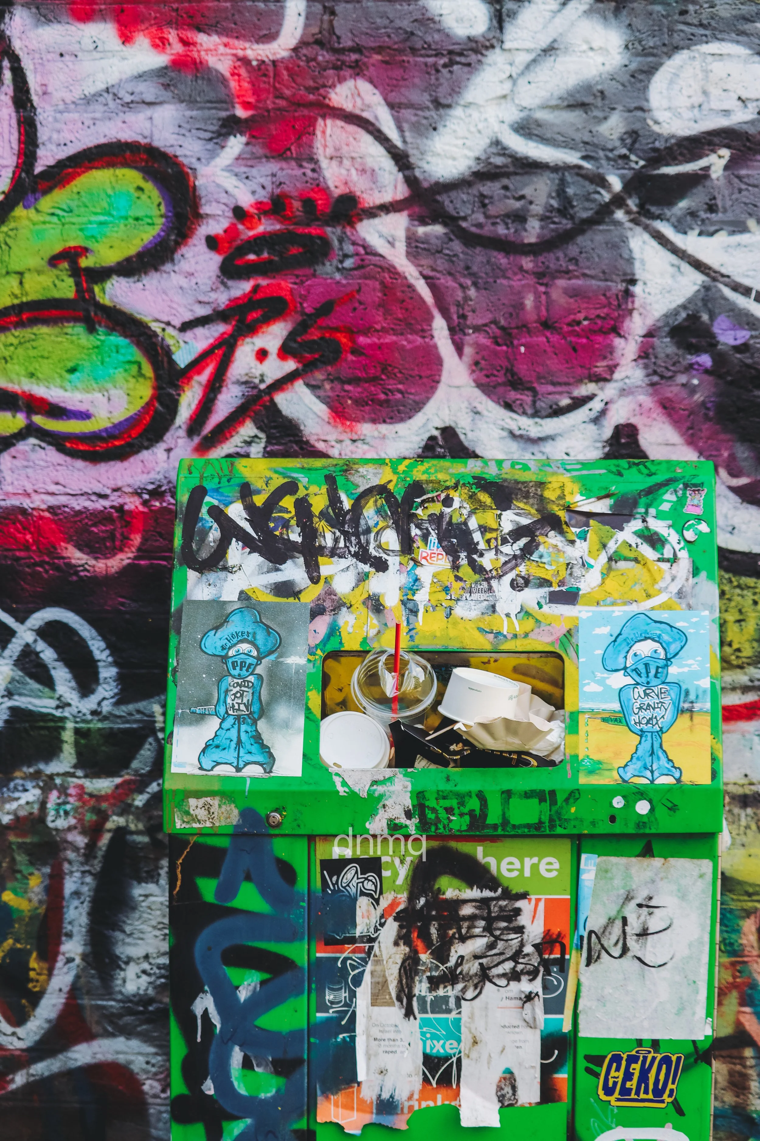 A green mailbox covered with graffiti, stickers, and posters is against a wall with colorful graffiti art.