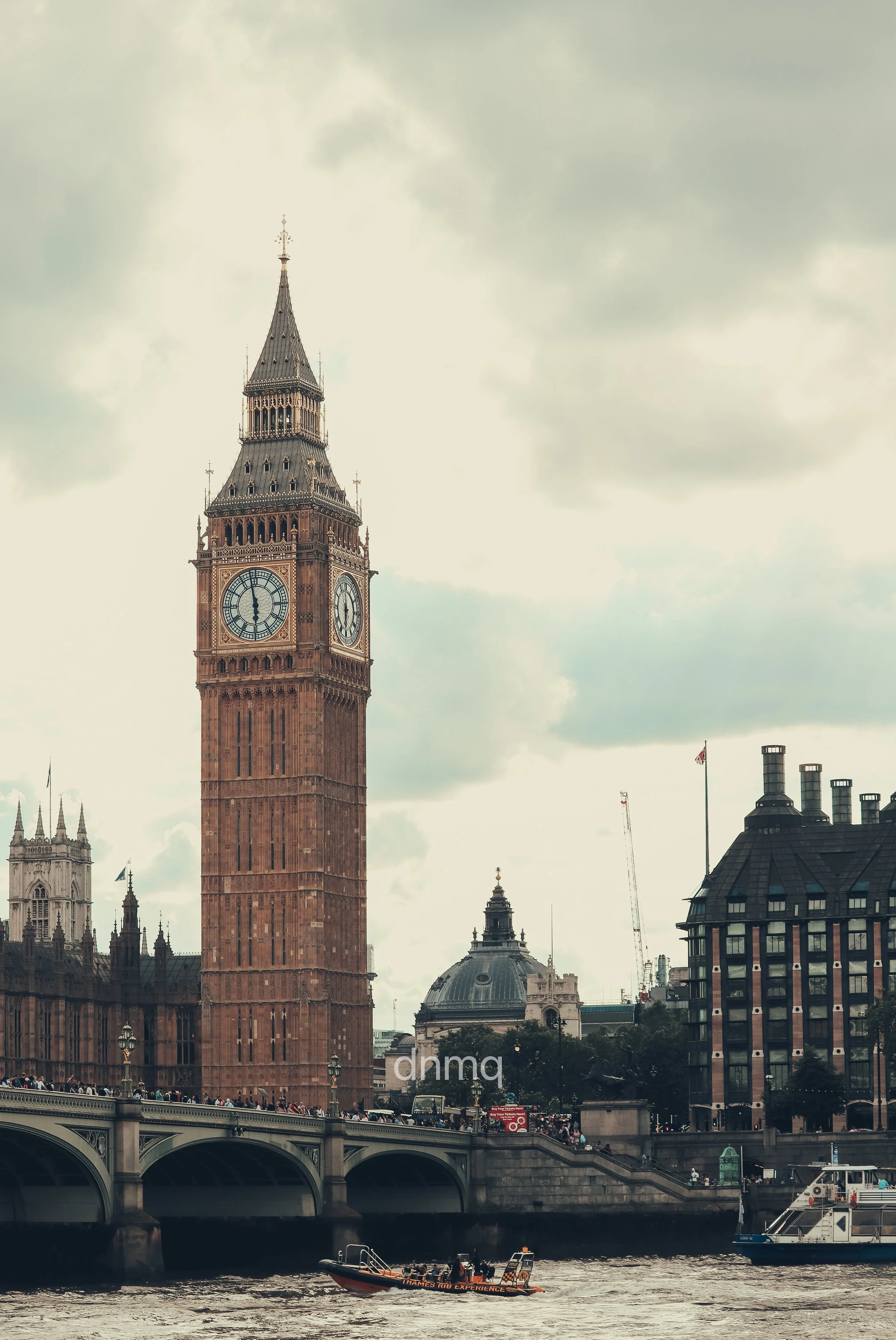 The image shows the Big Ben clock tower in London, with a bridge crossing a river below. Several boats are on the river, and there are buildings and cloudy skies in the background.