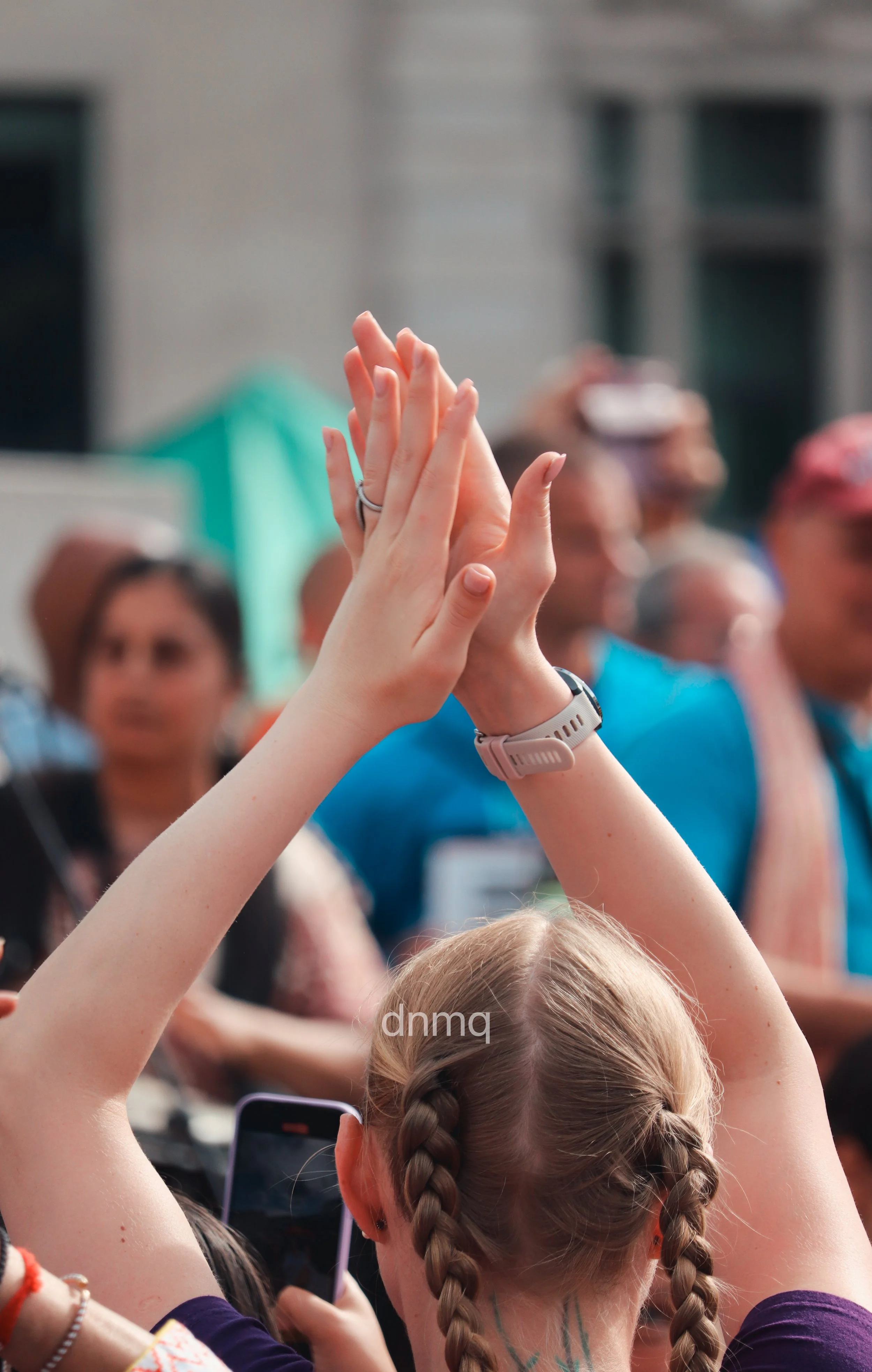 A young girl with braided hair is raising her hands middle-up in a high-five gesture in a crowd. Several people are blurred in the background, some holding phones and taking photos at an outdoor event.