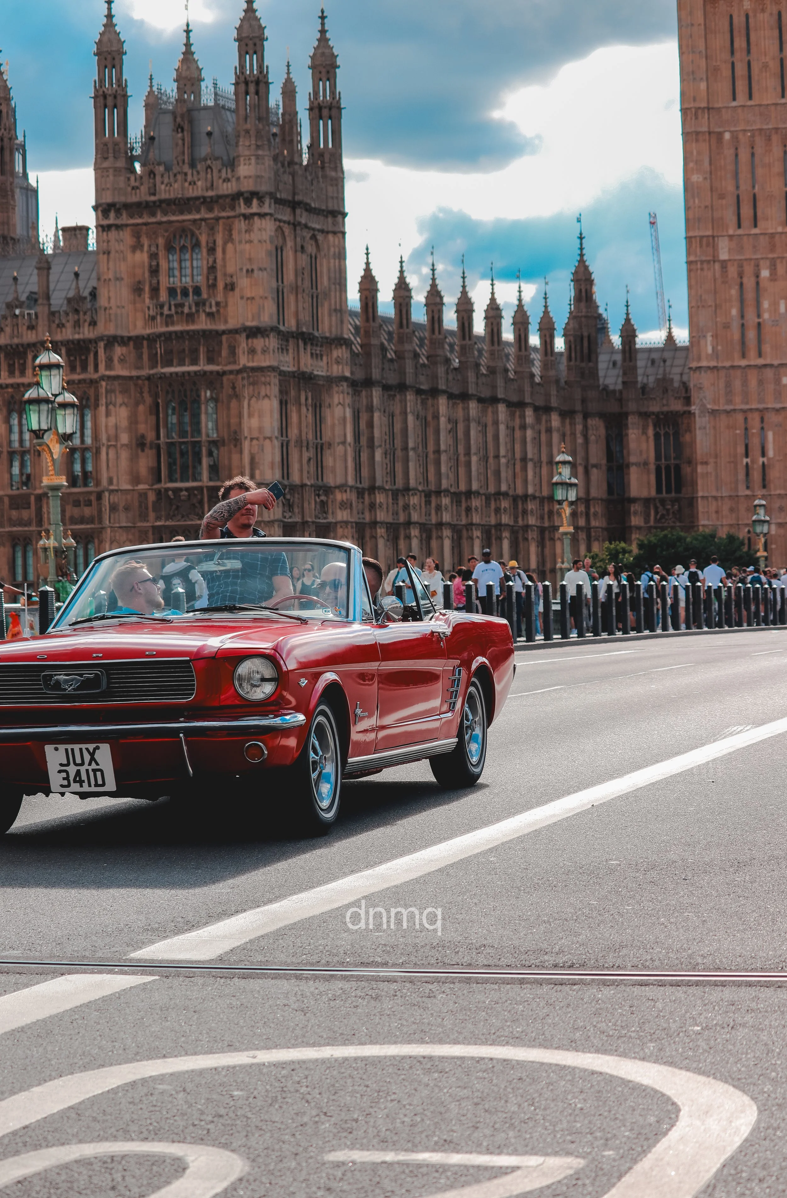 A red classic Ford Mustang convertible driving past the Palace of Westminster in London with onlookers and a cloudy sky.