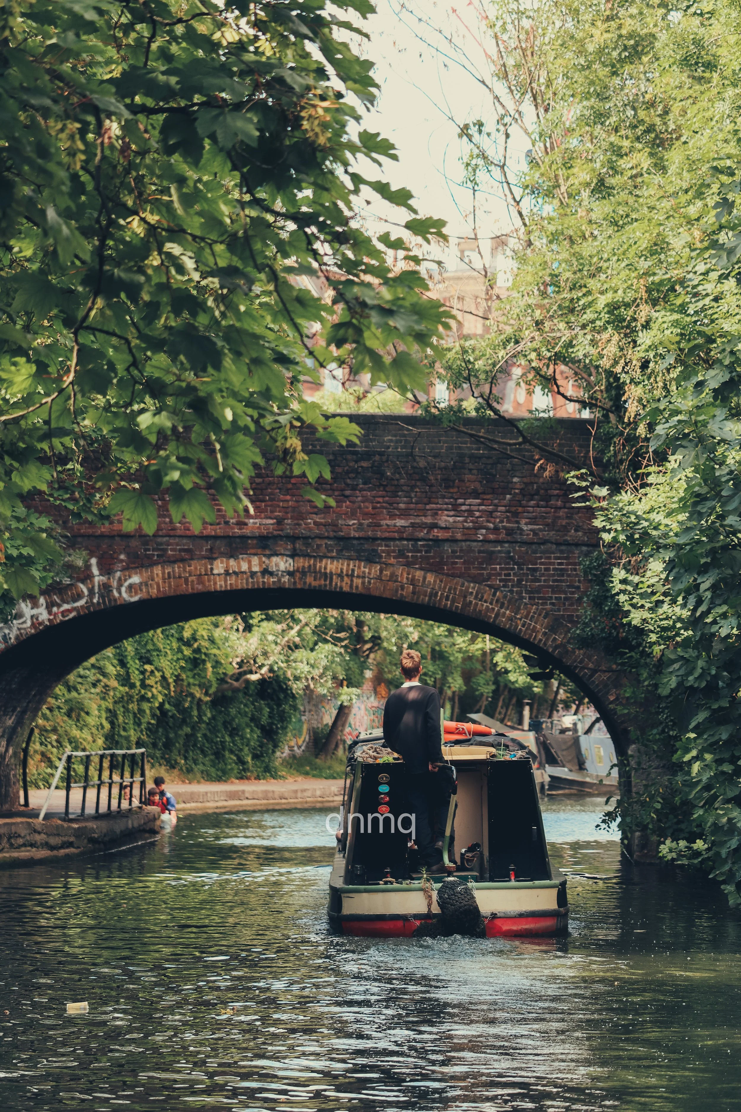 A boat with a person standing at the back is moving under a brick arch bridge on a narrow canal, surrounded by lush green trees.