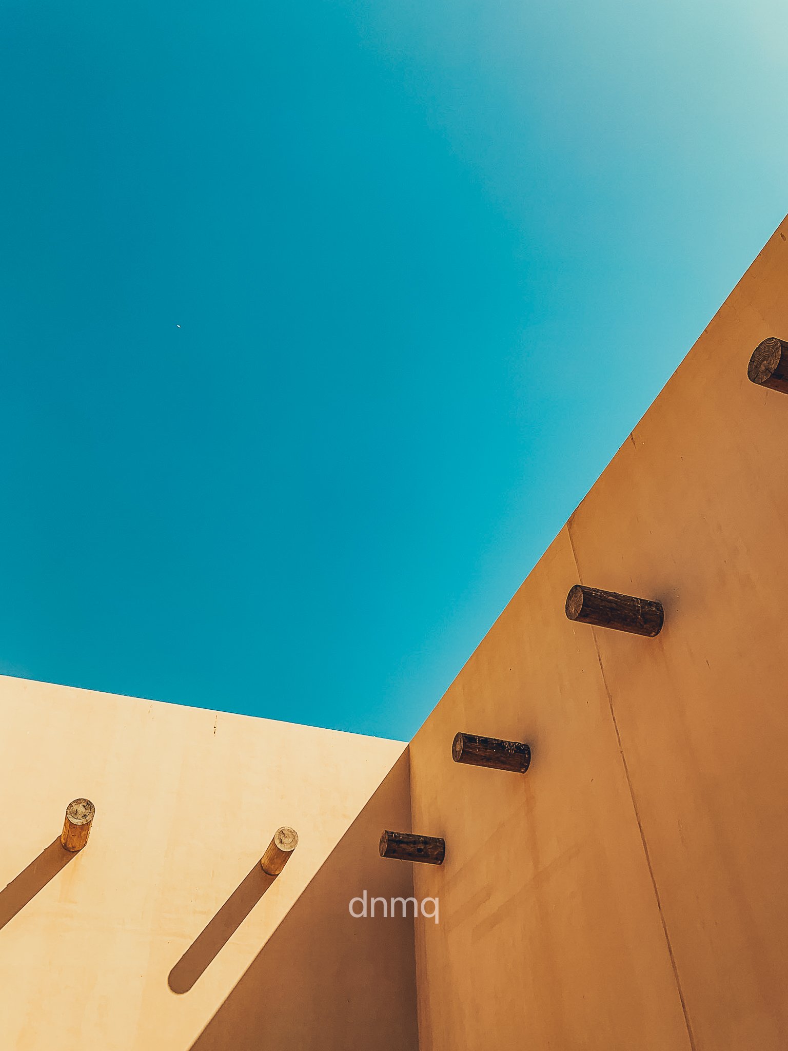 Minimalist photograph of a building corner with wooden beams protruding from the beige wall, against a bright blue sky with a small white cloud.