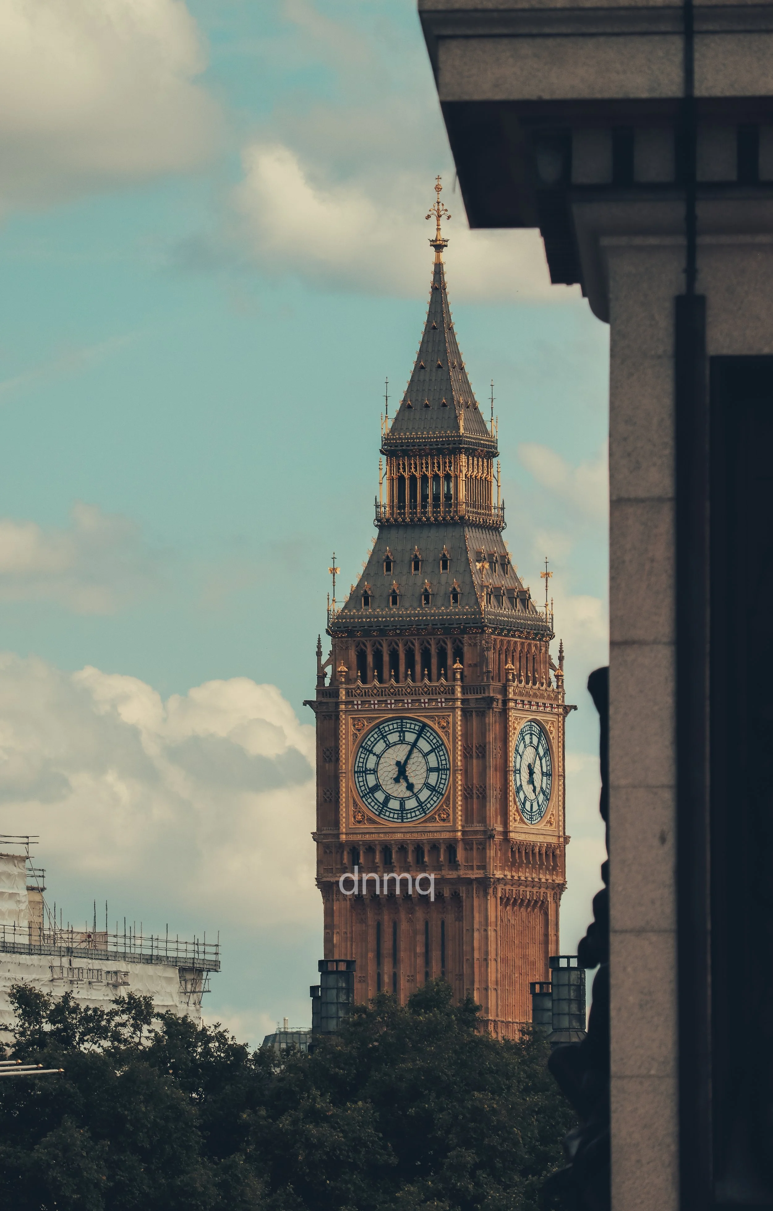 The clock tower of Big Ben, a landmark in London, England, with a partly cloudy sky background and green trees at its base, partially framed by a building on the right.
