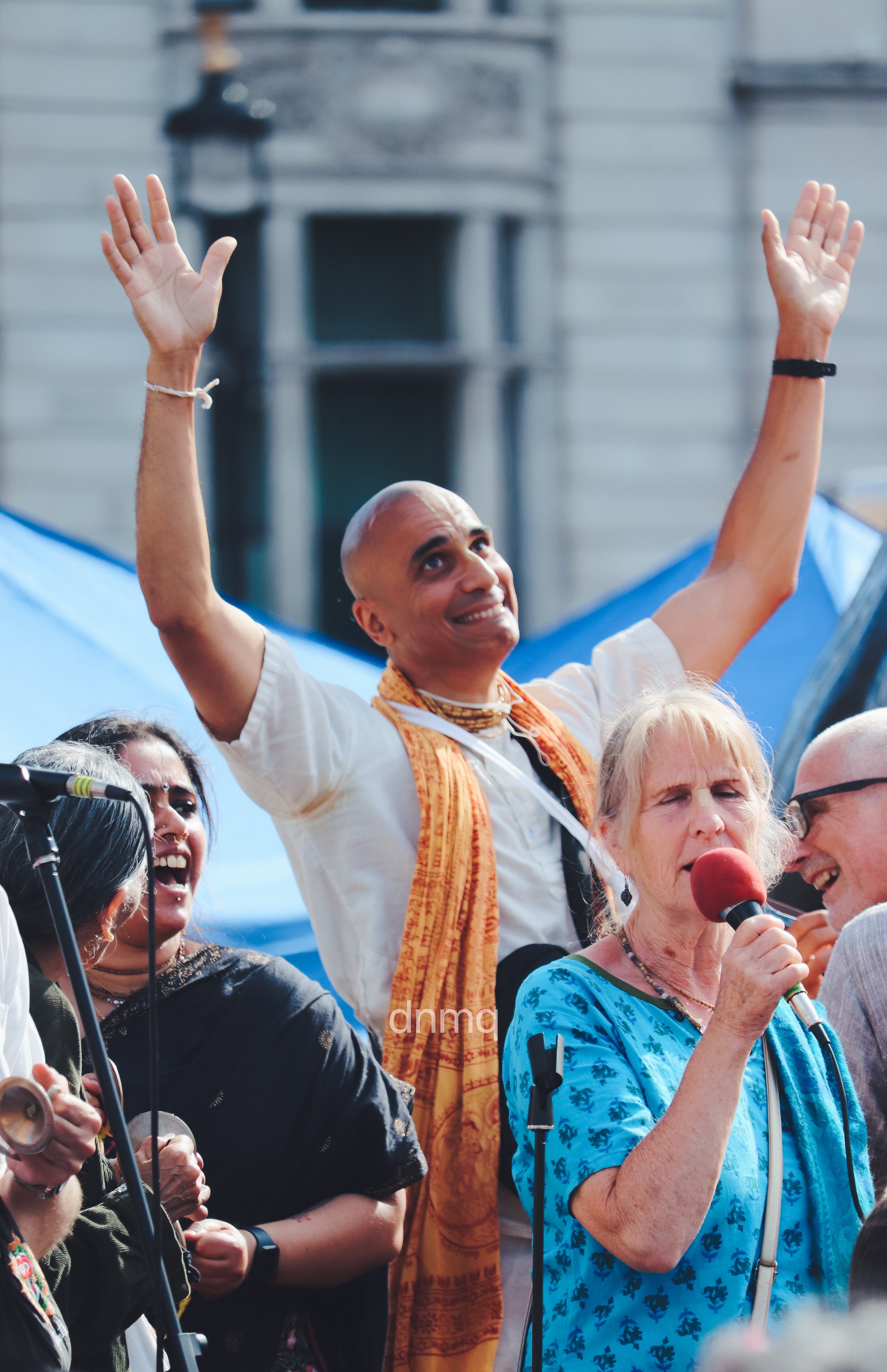 A diverse group of people at an outdoor event, one man with arms raised smiling, a woman with a microphone speaking, others laughing and enjoying.