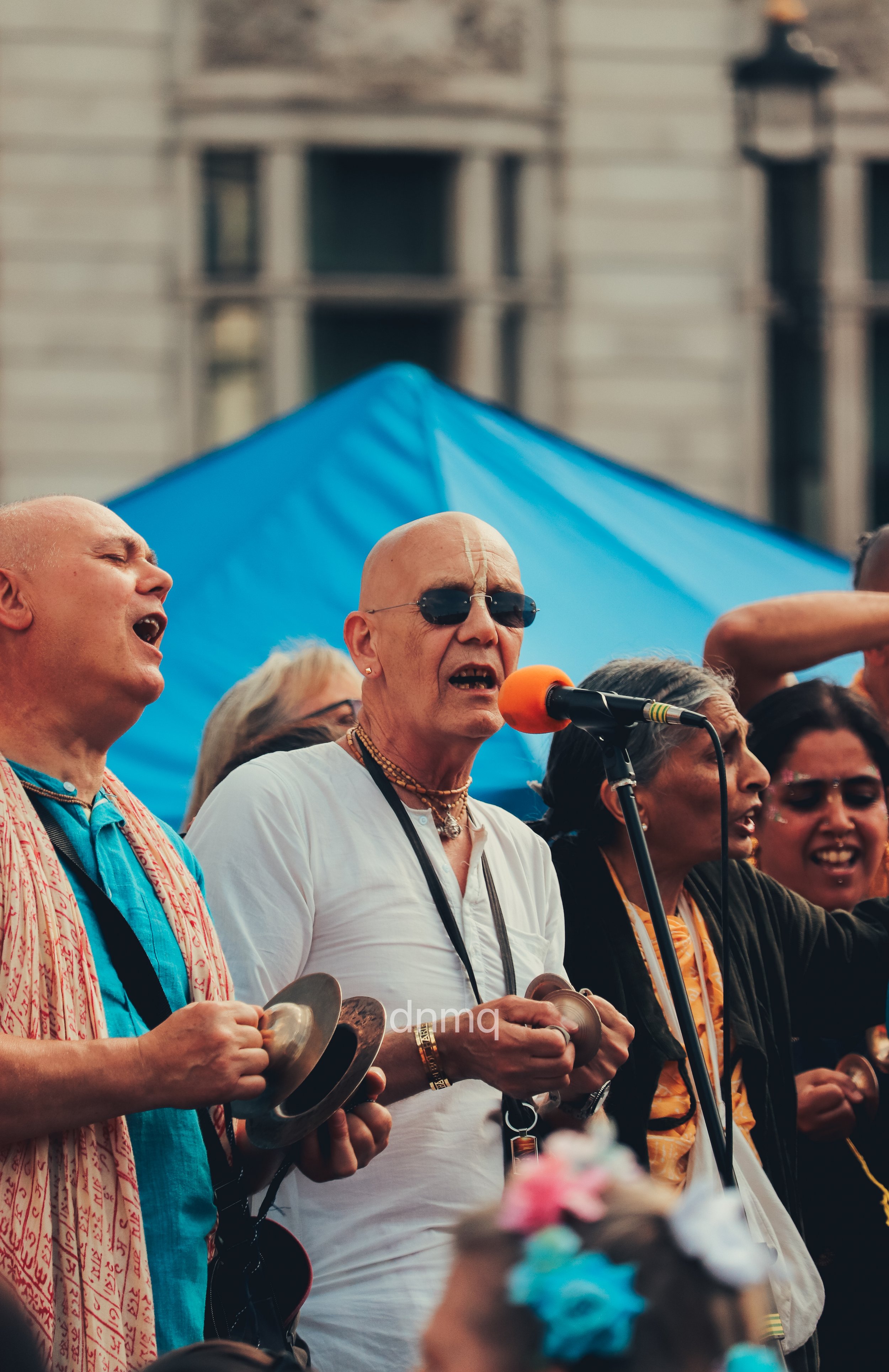 A group of people singing and playing musical instruments at an outdoor event, with a blue canopy and a historic building in the background.