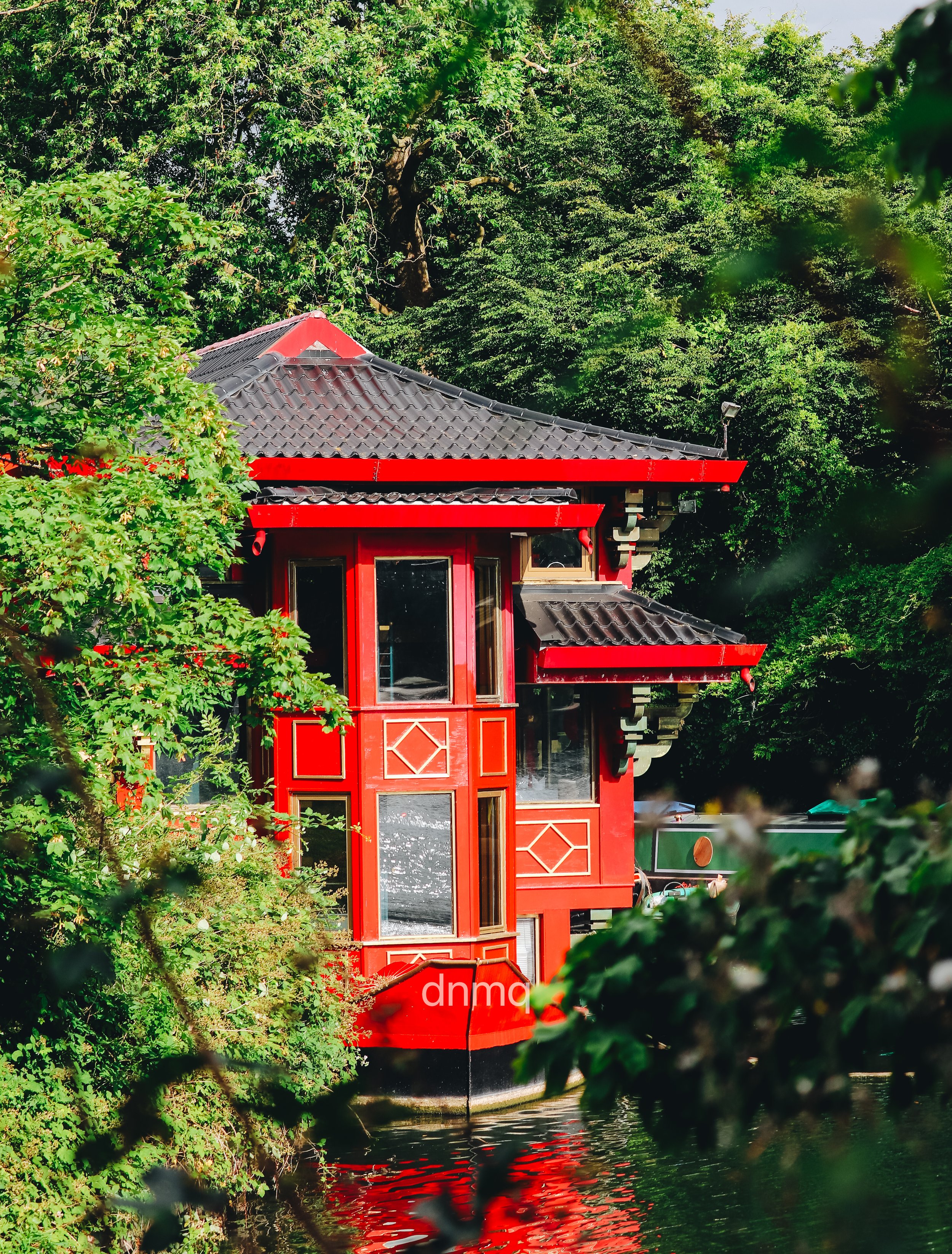 A red building with traditional Asian architectural features, including a tiled roof and wooden framework, is situated next to a body of water and surrounded by lush green trees.