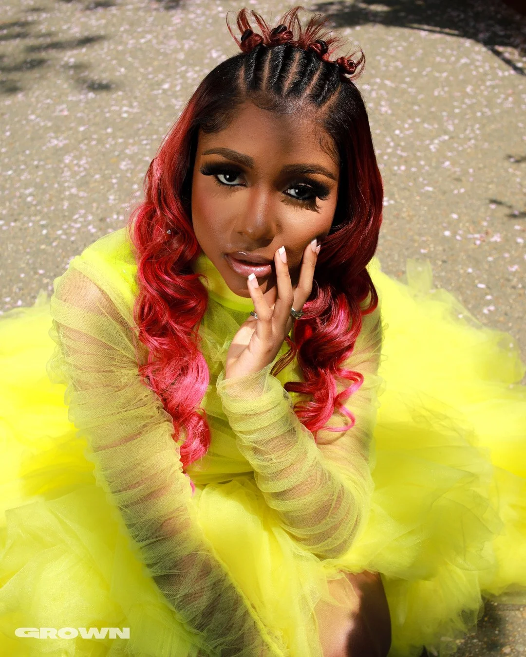 A woman with pink and black hair styled in curly waves, wearing a yellow sheer long-sleeve top and a fluffy yellow skirt, sitting on the ground covered with fallen flower petals.
