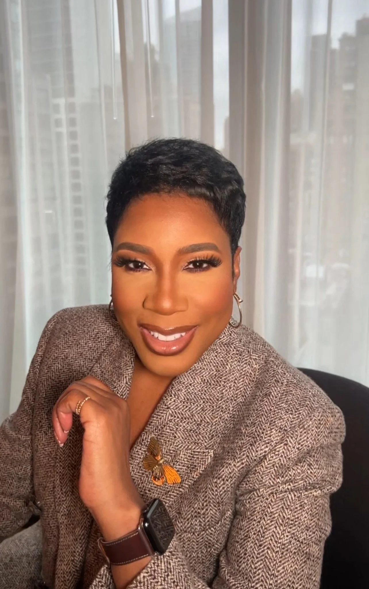 A woman with short black hair, wearing a brown checked blazer with a butterfly pin, a smartwatch, and gold earrings, sitting in front of a window with sheer white curtains, smiling at the camera.