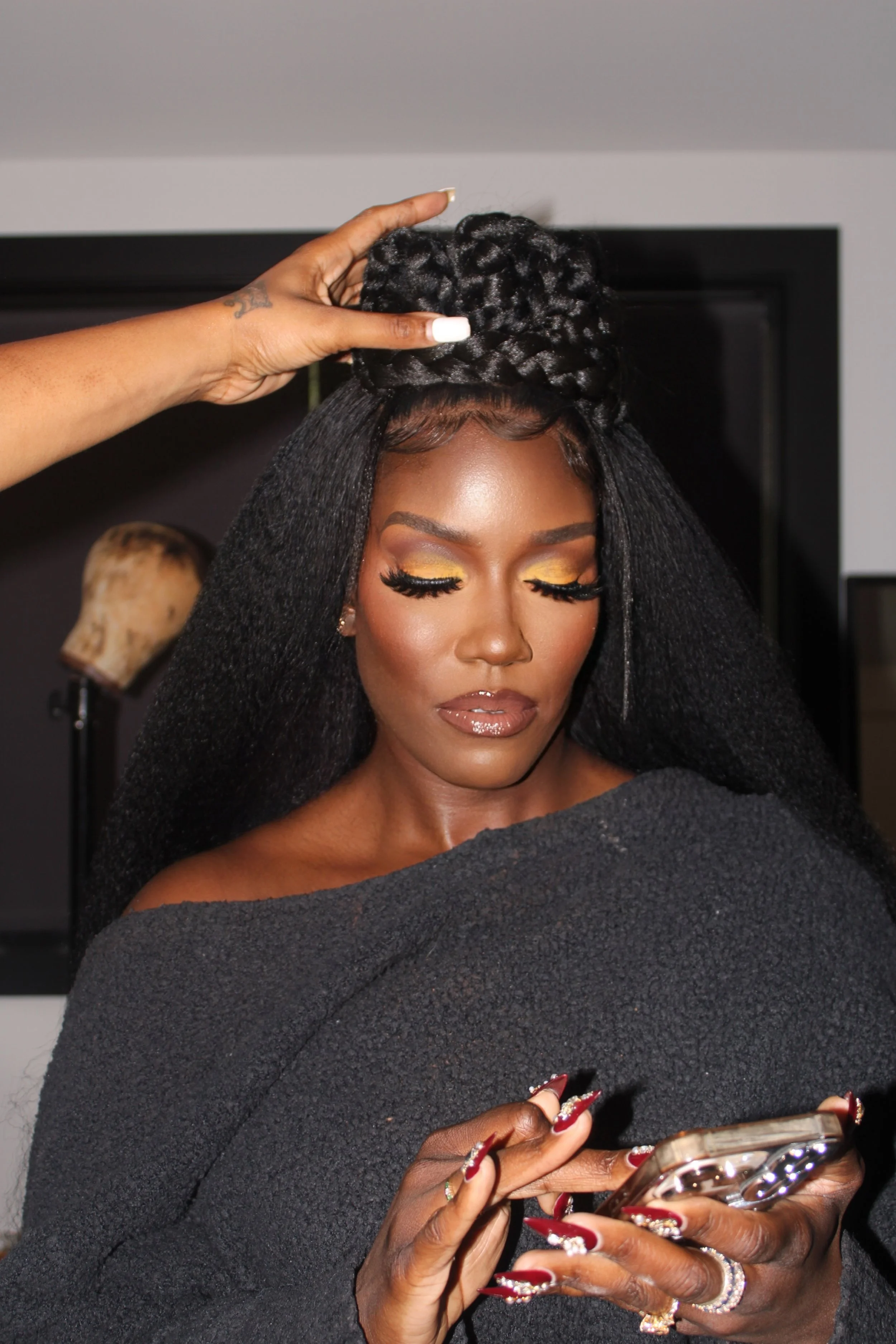 A woman with long black hair styled with a braided high bun, wearing makeup with yellow eyeshadow, and looking at her phone while a stylist adjusts her hairstyle.