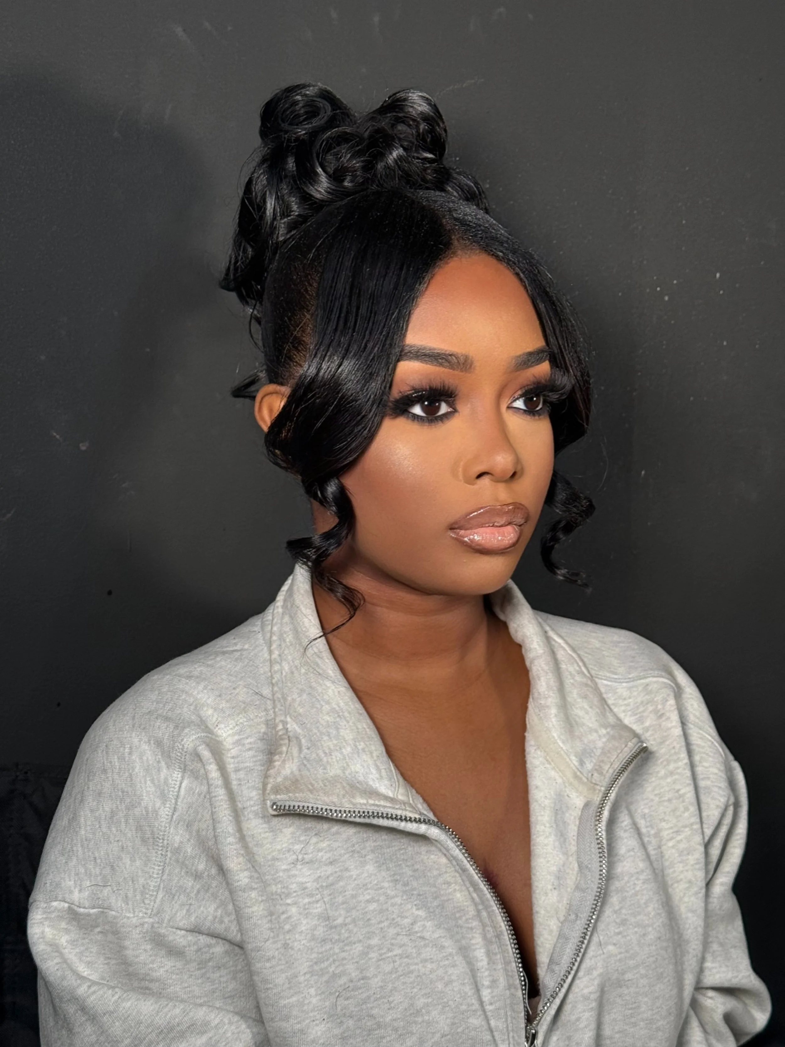 A woman with dark hair styled with curls, wearing makeup, and dressed in a light gray zip-up sweatshirt, posing against a black background.