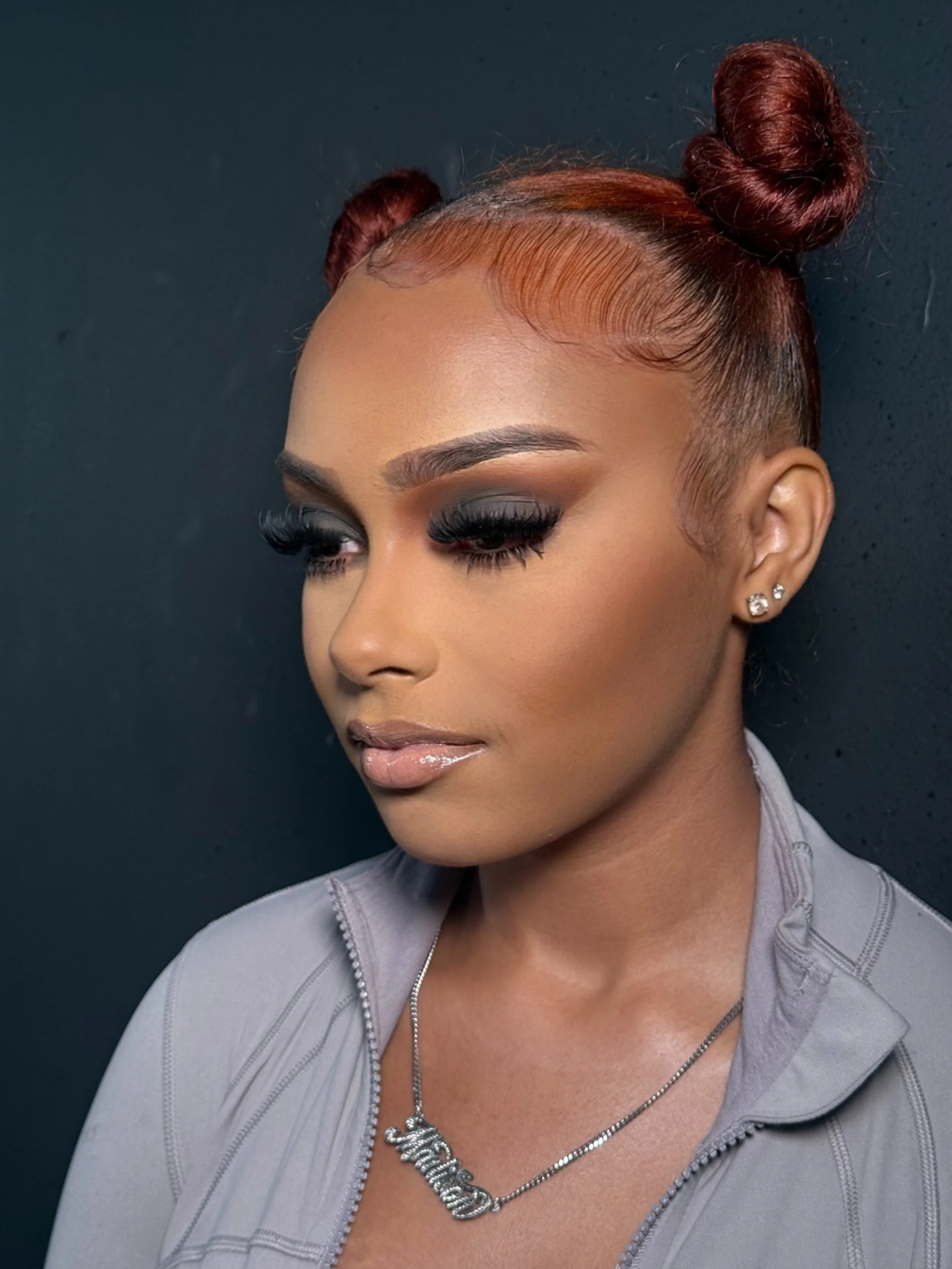 A woman with a light brown complexion and red hair styled in two buns, wearing makeup with dark eye shadow and glossy lips, gold earrings, a silver necklace with a nameplate, and a light gray zip-up top, standing against a dark background.