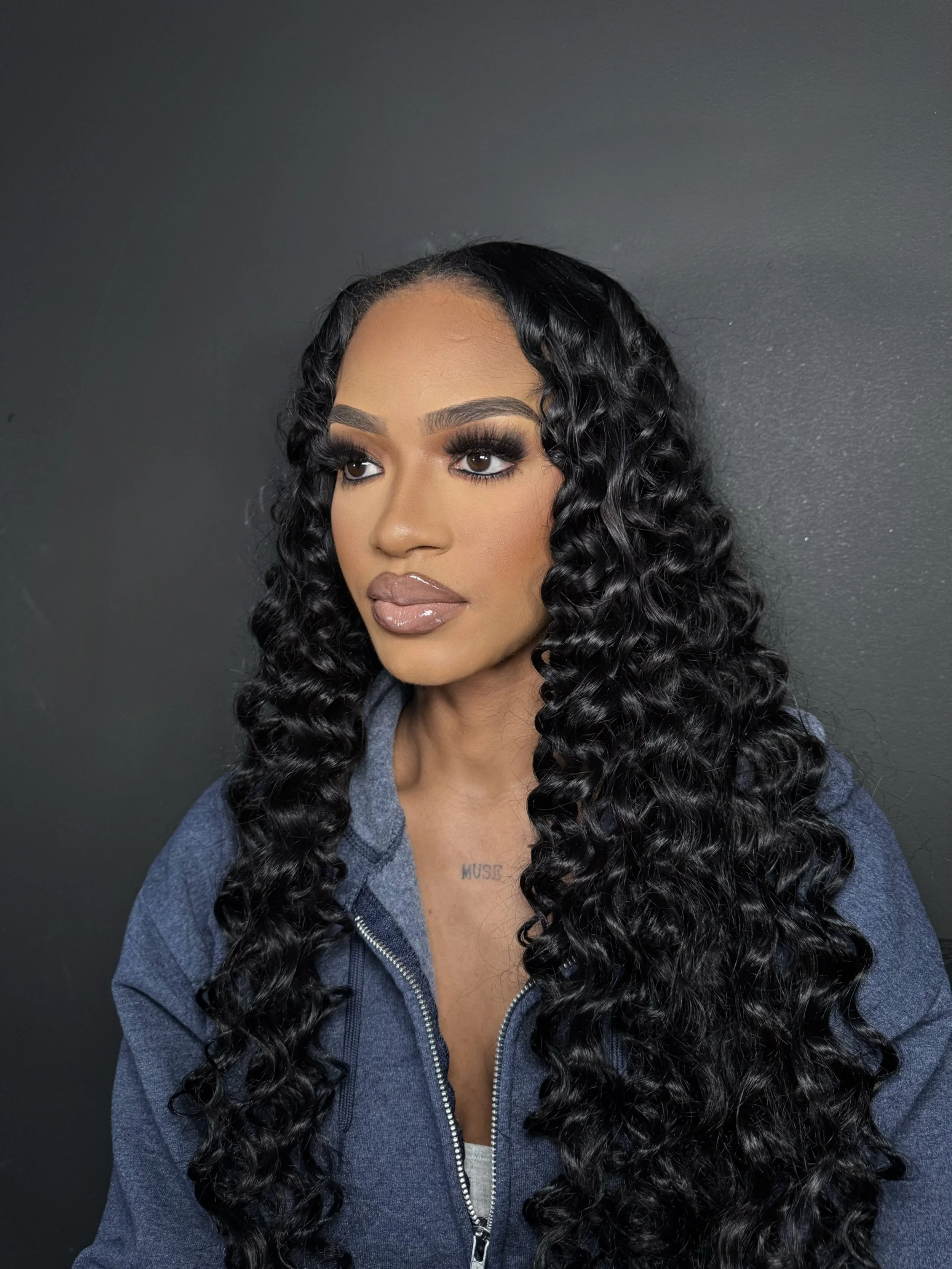 A woman with long, curly black hair and makeup, wearing a blue jacket, against a dark gray background.